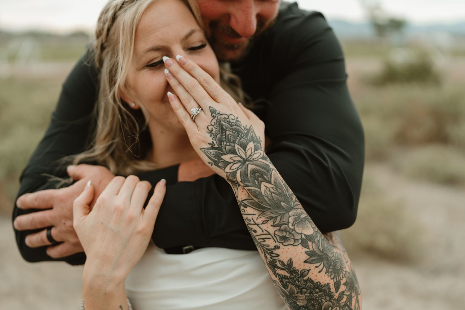 Couple embracing outdoors, woman with floral sleeve tattoo laughing, man in black shirt hugging her from behind.