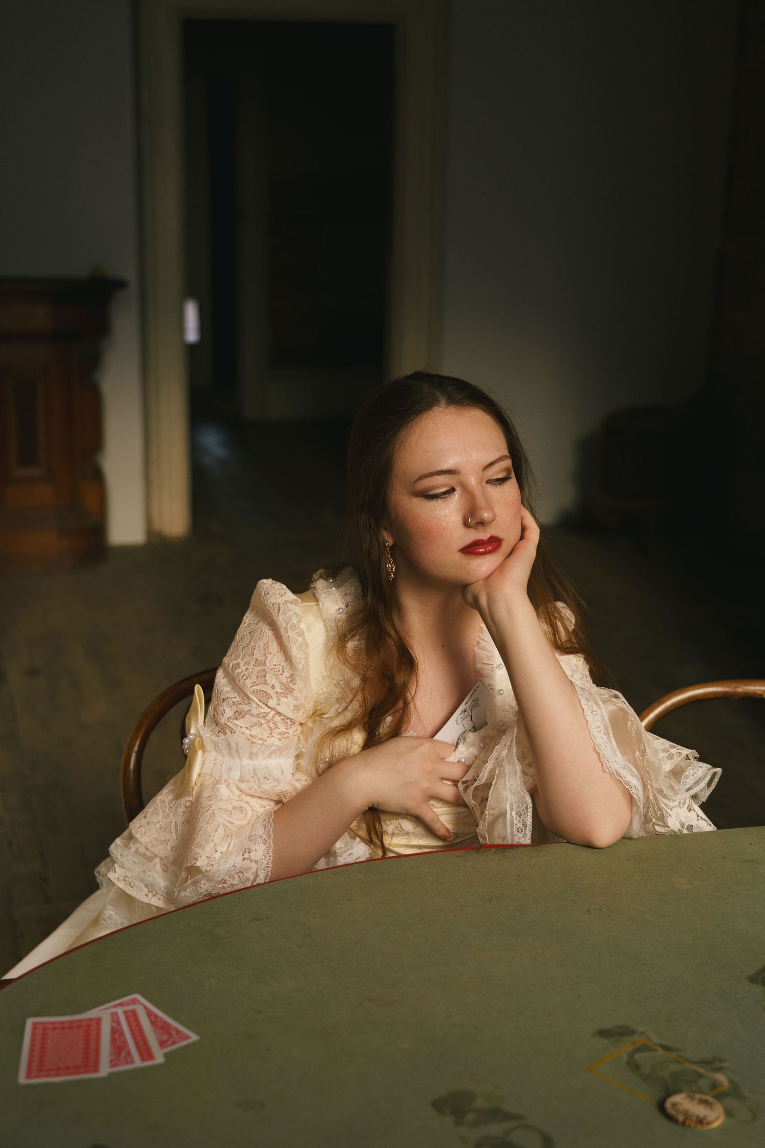 Woman in vintage lace dress sitting at green table with playing cards, looking pensive in moody light.