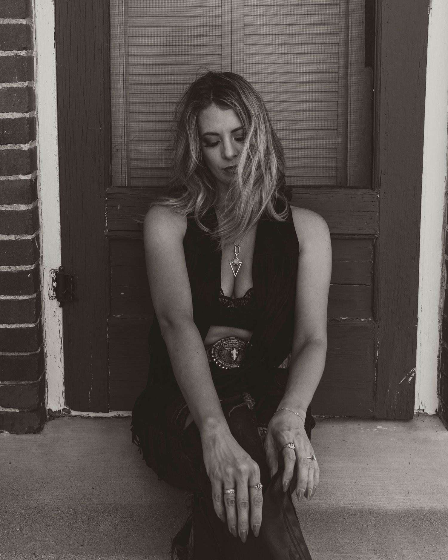 Moody black and white portrait showing a person in a black outfit seated by a door with shutters.