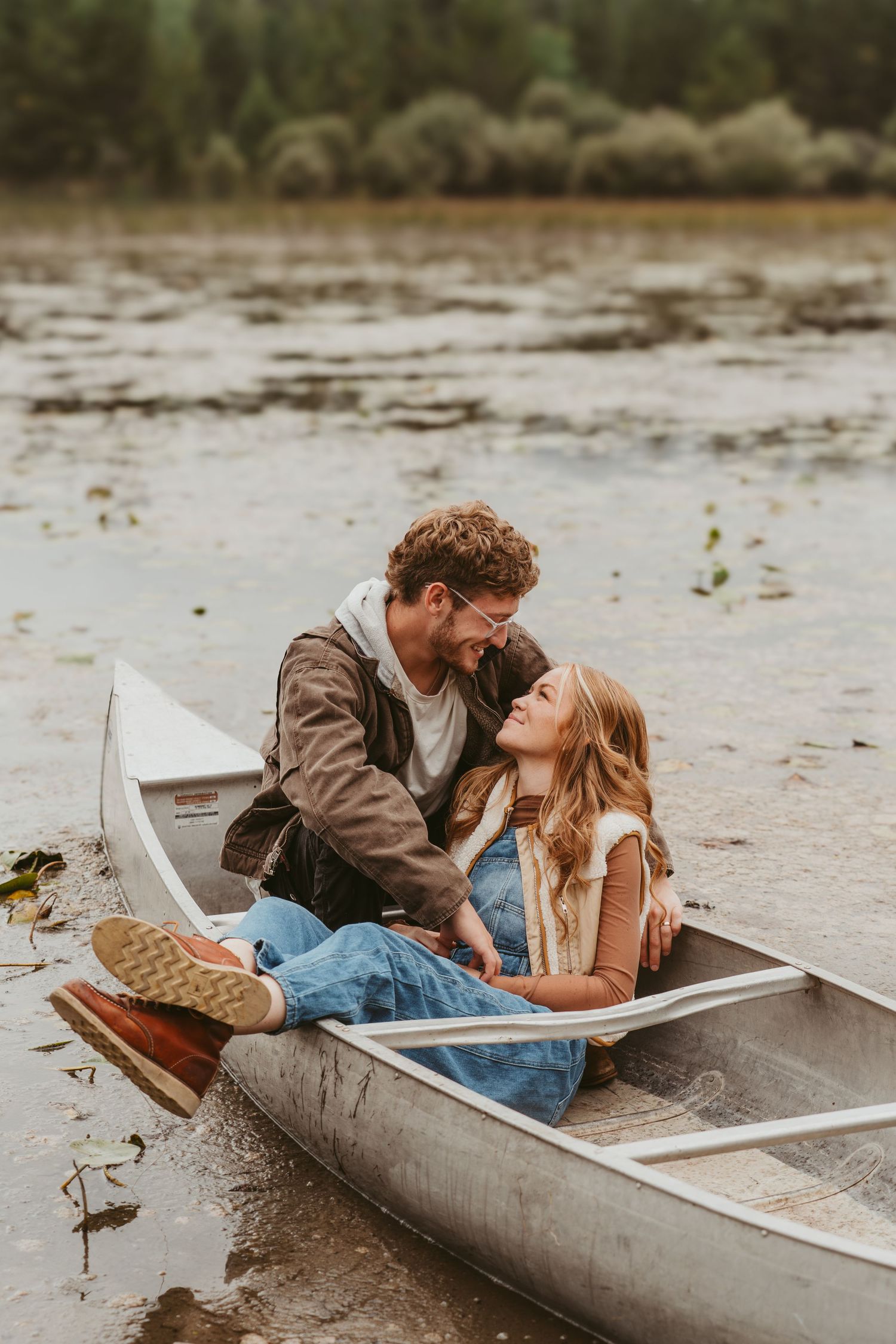 A couple shares an intimate moment while laying in a silver canoe on a cloudy day at the lake.
