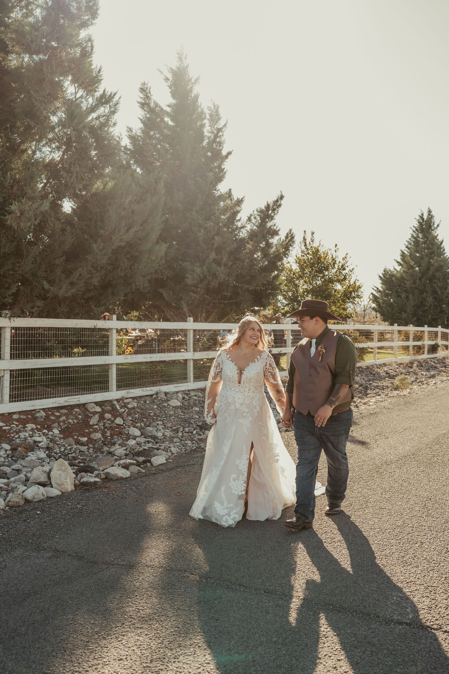 A couple walks together on a sunlit path with white fencing and evergreen trees in the background.