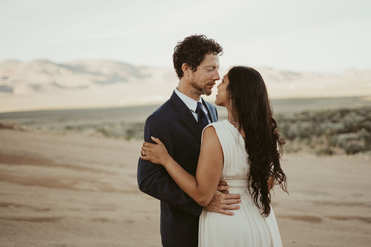 A couple shares a romantic embrace in a desert landscape, the groom in a navy suit and bride in a white dress.