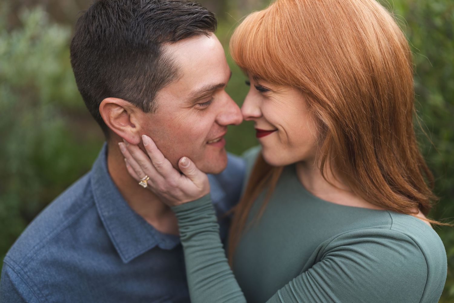 A couple shares a romantic moment outdoors, touching foreheads and smiling, the woman with red hair gently holding the mans face.