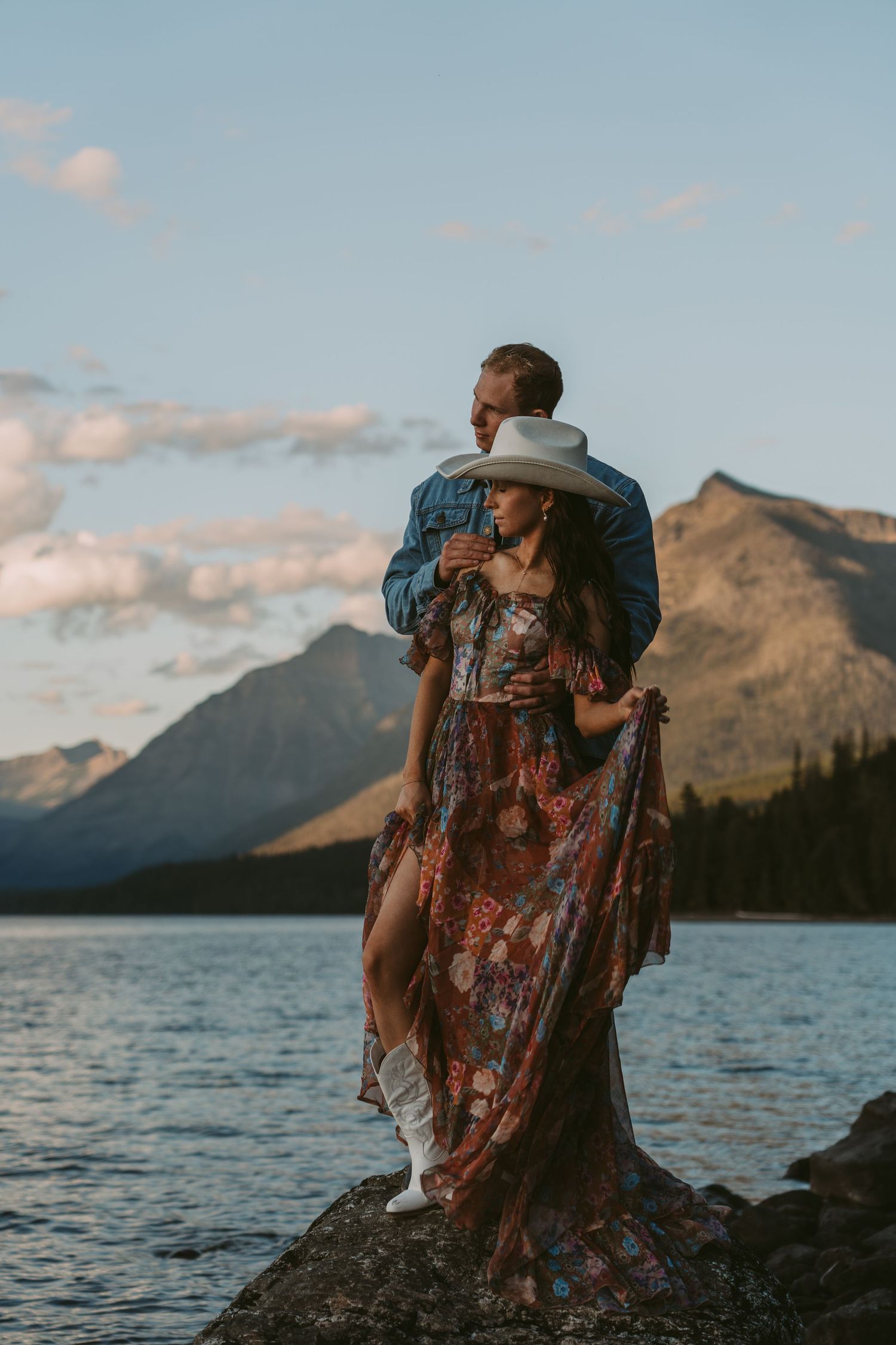 A couple sits together on rocky shores of a mountain lake at sunset, embracing against a backdrop of majestic peaks.