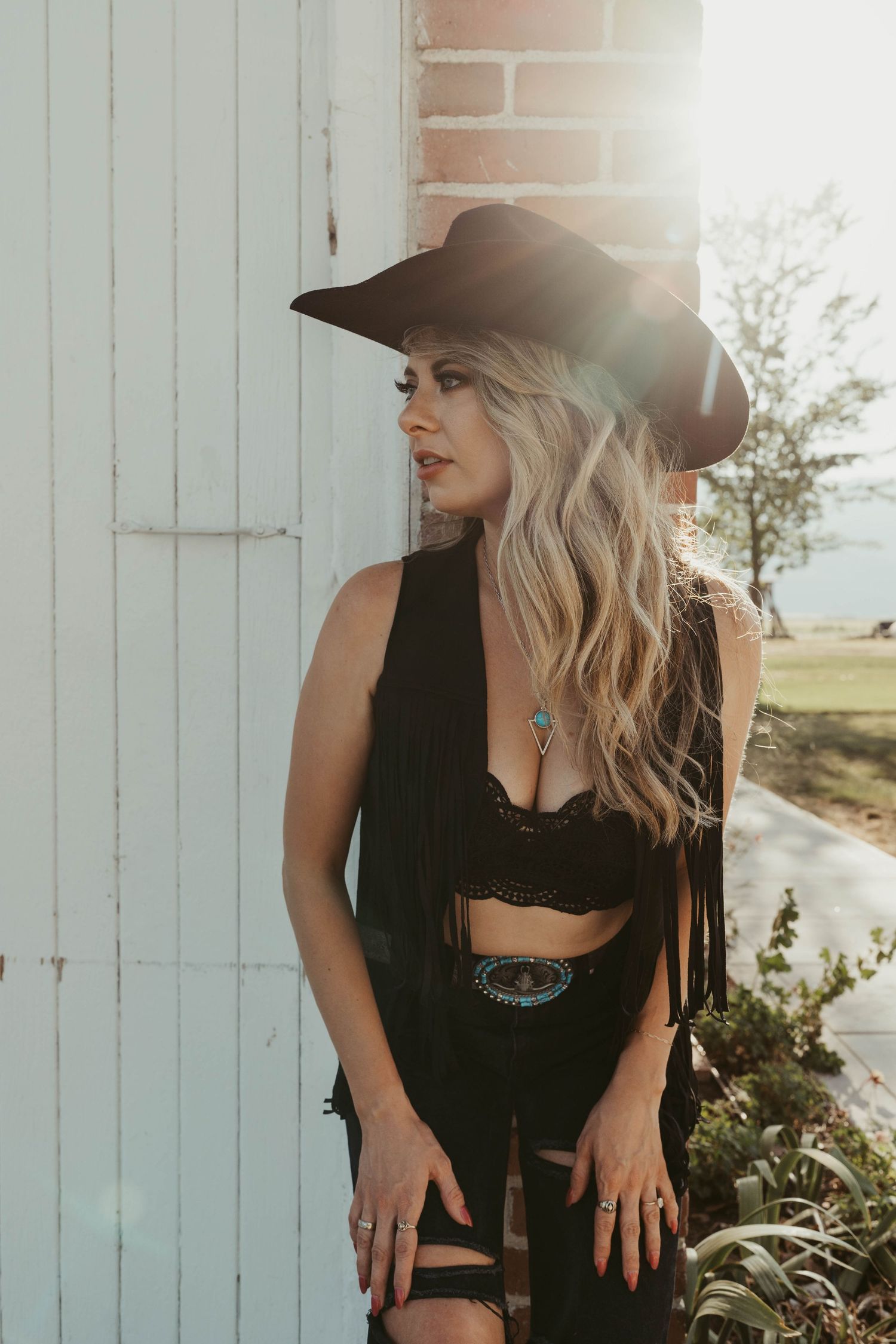 Woman in black outfit and hat poses against rustic white wooden wall during golden hour photoshoot.