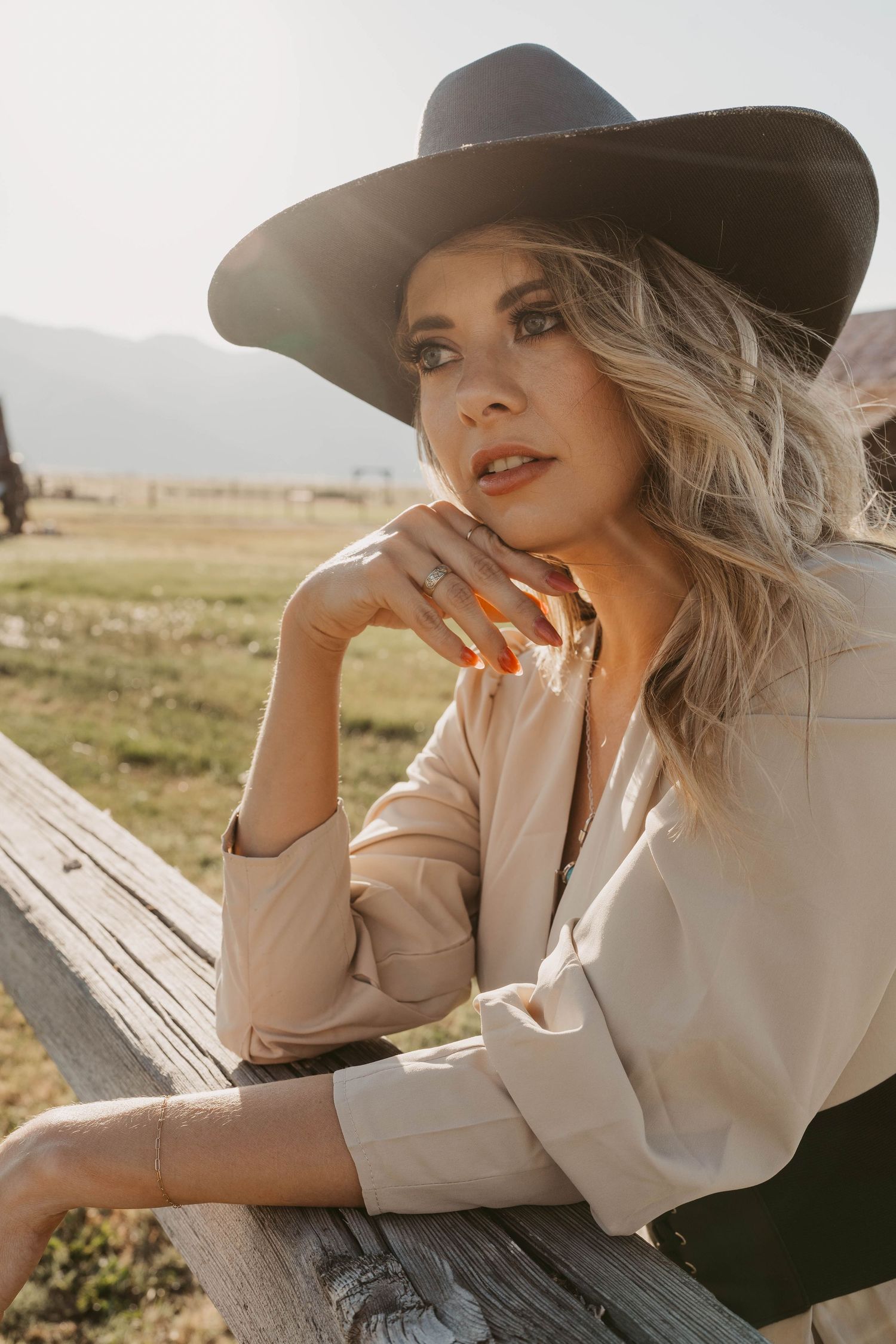 Sun-drenched outdoor portrait of someone with blonde hair wearing a tan dress and wide brimmed hat while resting on wooden fence.