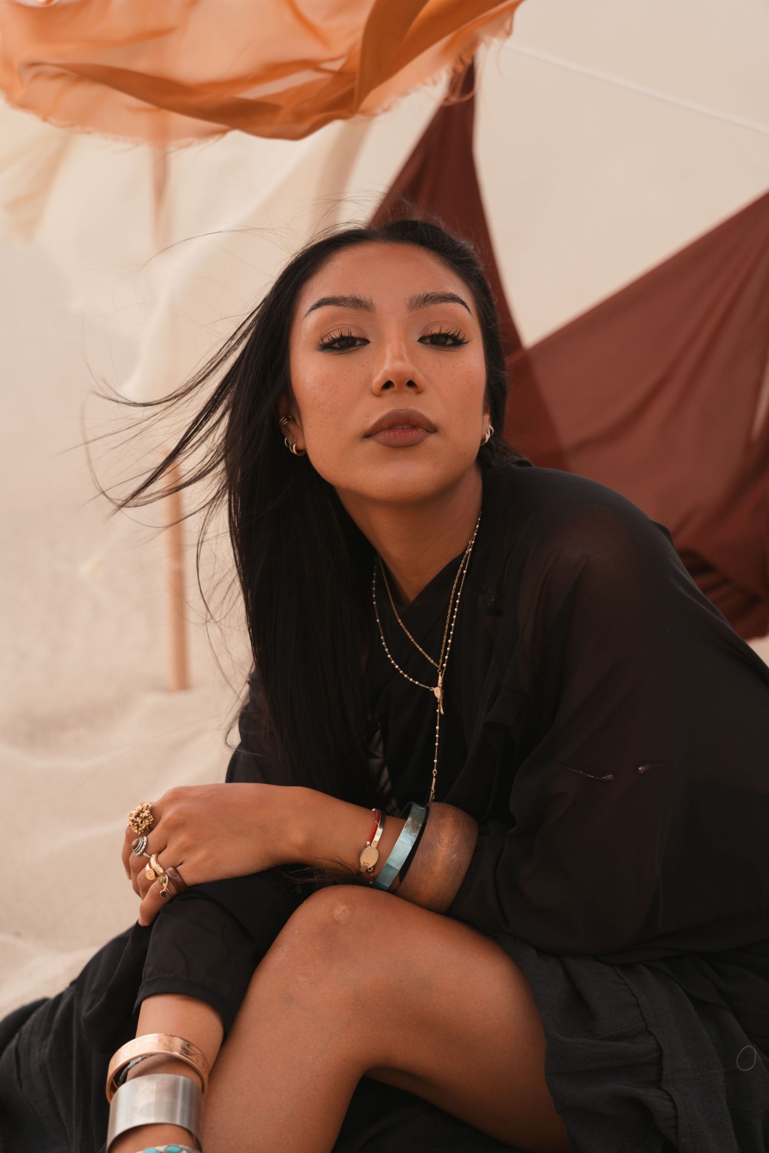 Woman in black outfit with gold jewelry and bracelets poses against colorful fabric backdrop.