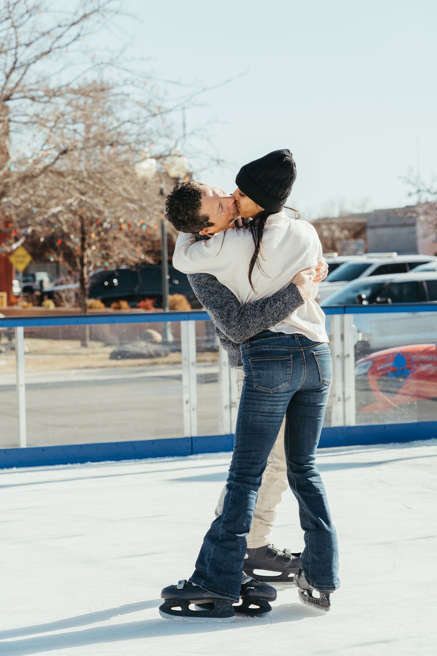 A couple embraces while ice skating on a snowy outdoor rink with parked cars and bare trees in the background.