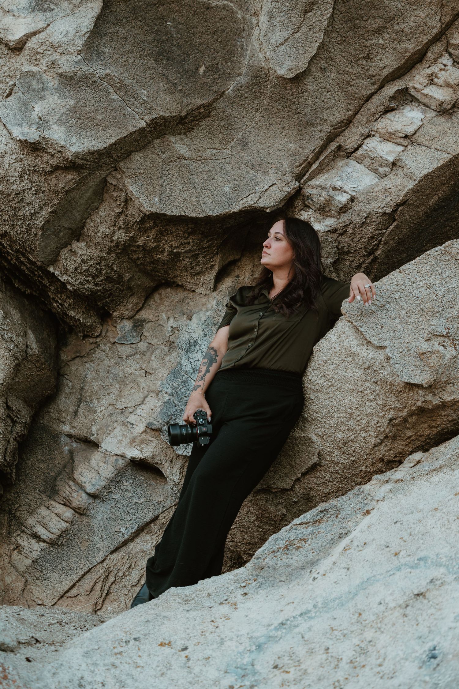 Woman in olive shirt and black pants holds camera while leaning against large rock formation outdoors.