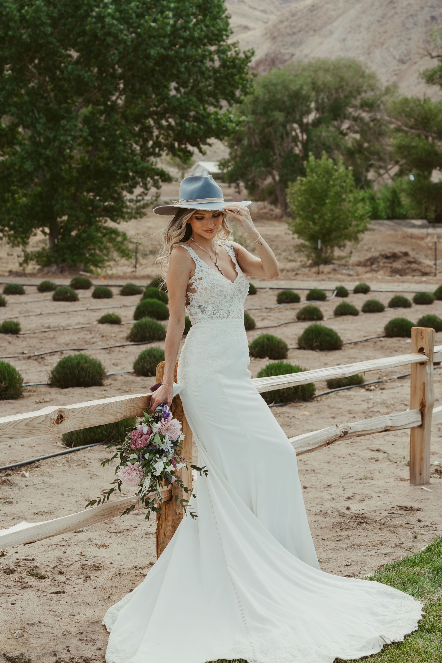 Bride in white lace gown and gray hat holds floral bouquet by rustic fence in desert landscape.
