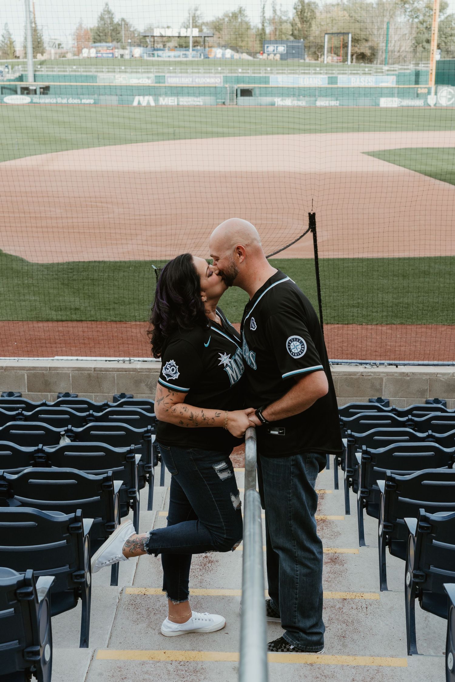 Couple sharing a kiss in baseball stadium stands, both wearing matching black jerseys, with the diamond field visible in background.