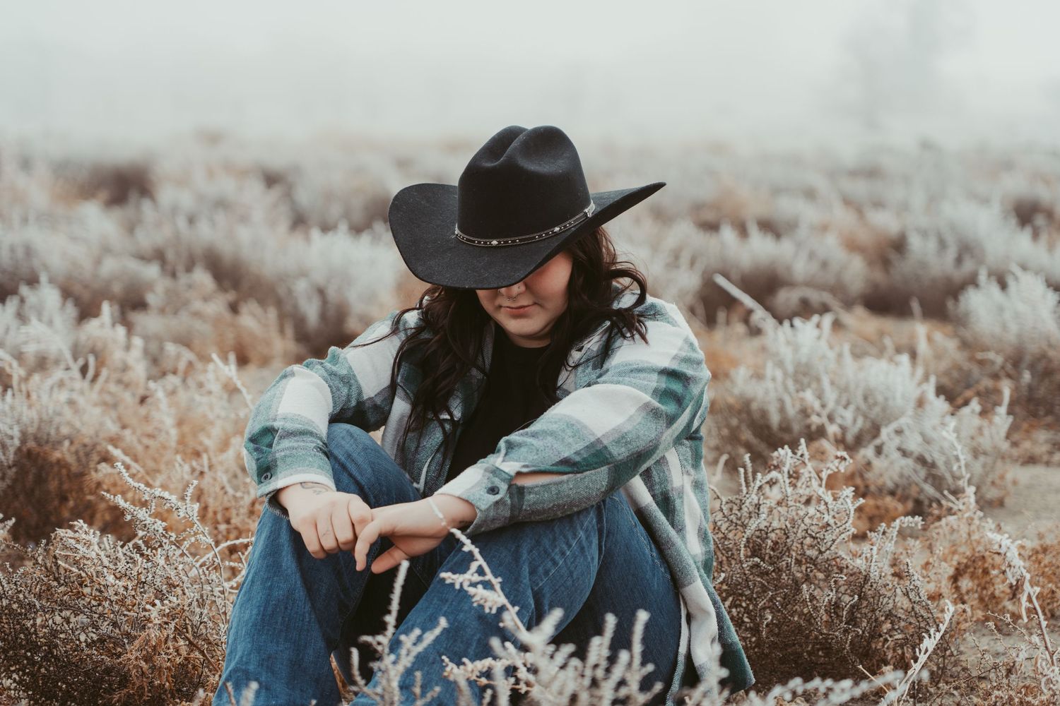 Someone in a black cowboy hat and denim jacket sits in a field of dried sagebrush on a moody overcast day.