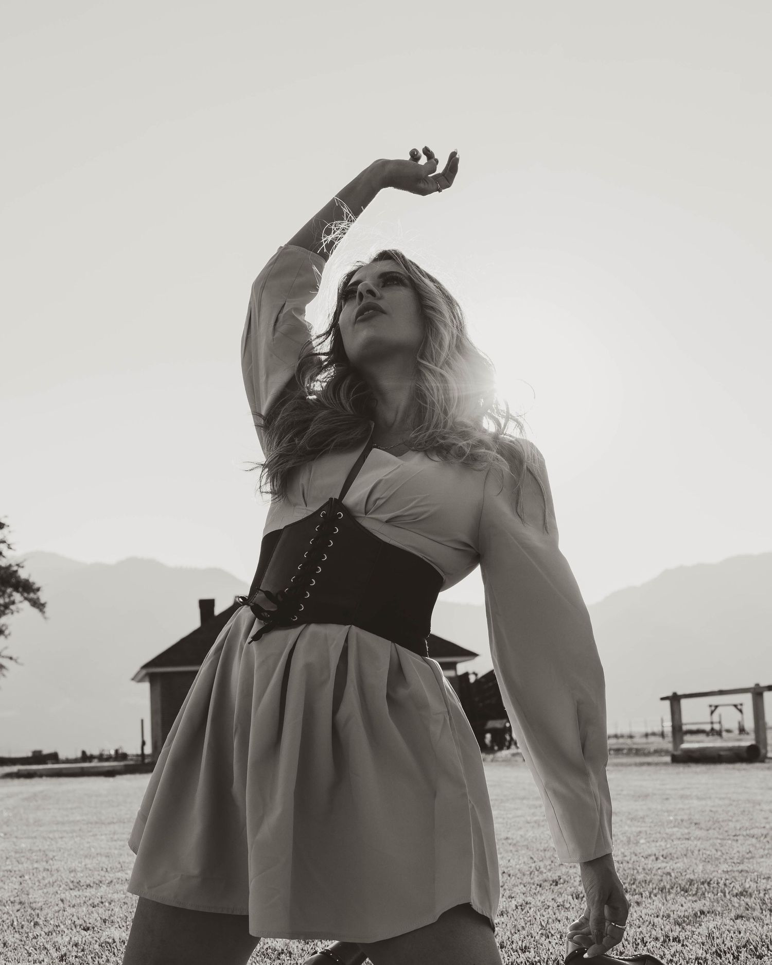 Artistic black and white photo of a figure in a corseted dress and long sleeves posing outdoors with mountains in background.