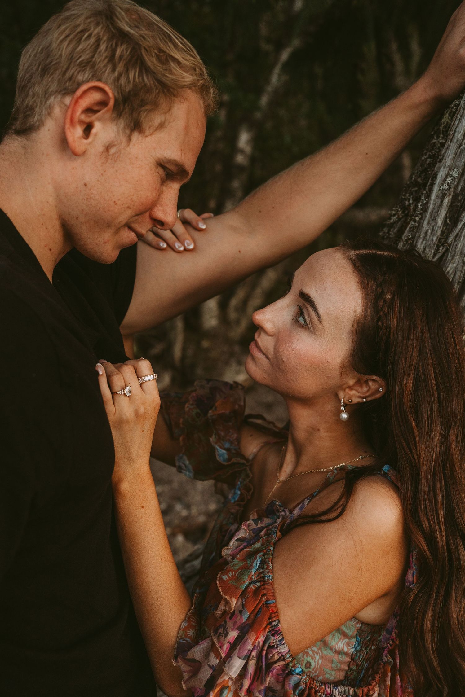 Romantic couple sharing intimate moments against a tree in moody forest lighting with warm earth tones.