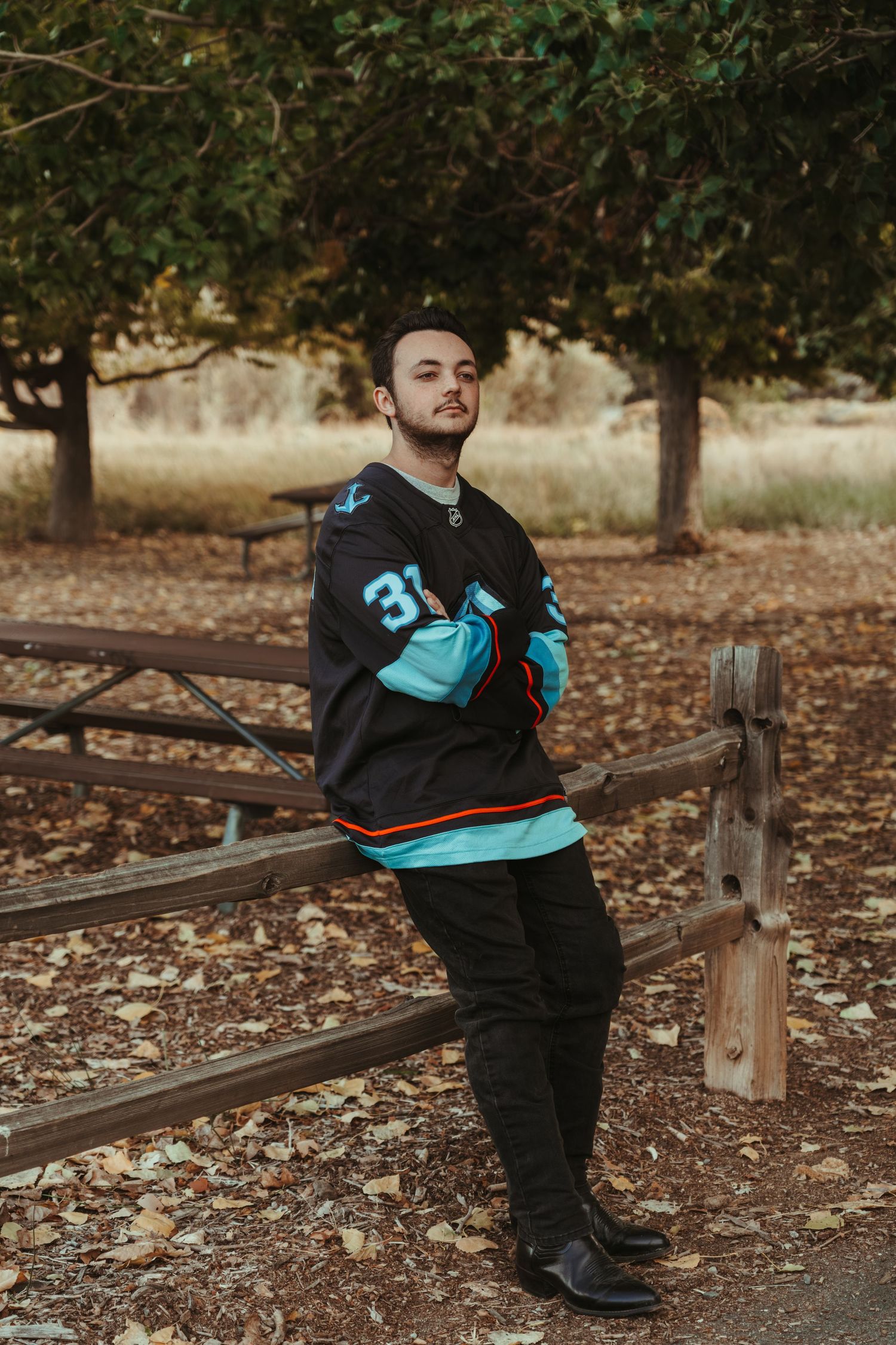 Person wearing black and turquoise jersey sits on rustic wooden fence in autumn setting.