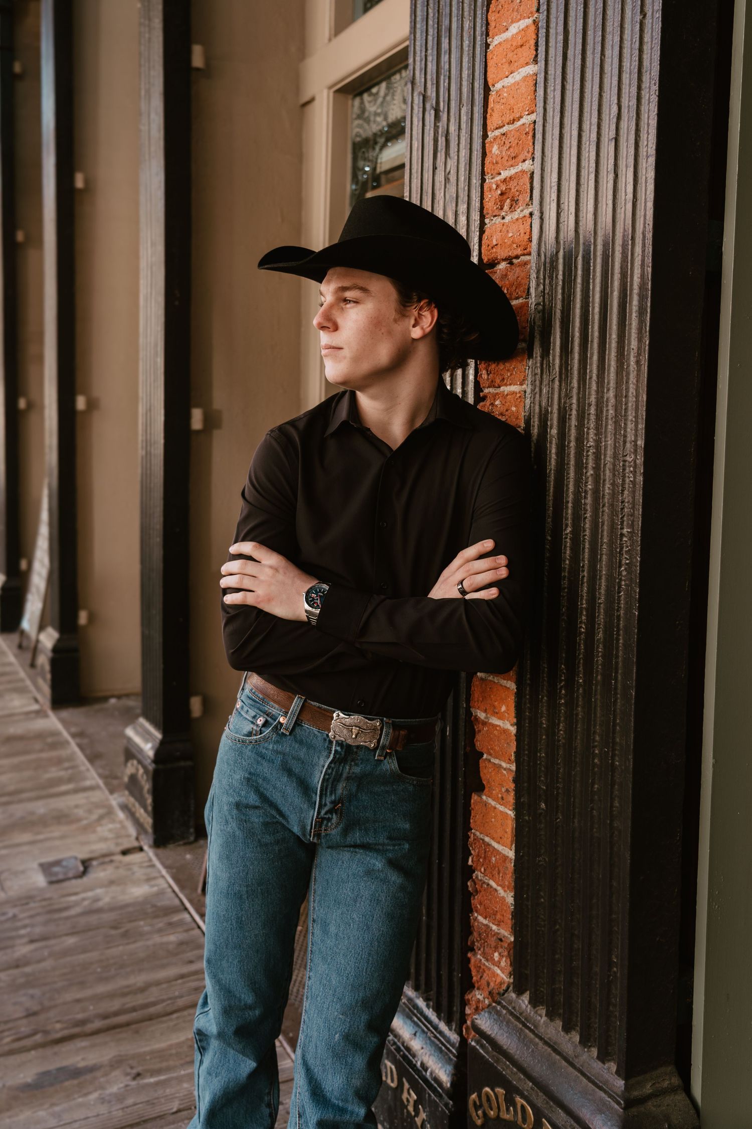 Person in black cowboy hat and denim leans against dark column in moody lighting.
