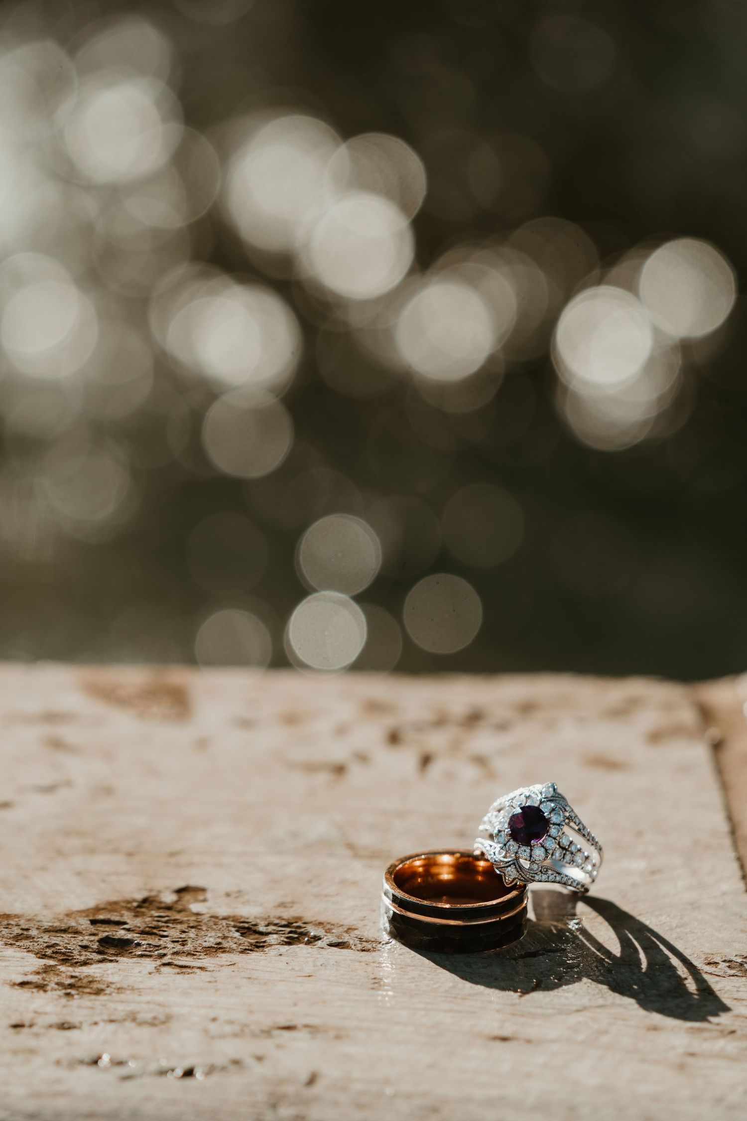 Wedding rings and diamond engagement ring casting shadows on a textured stone surface with bokeh background.