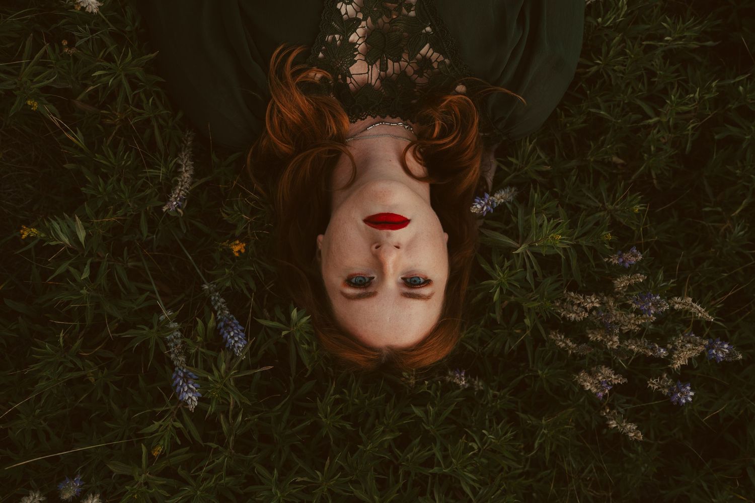 A portrait of a woman with red hair lying in a field of purple wildflowers at dusk.