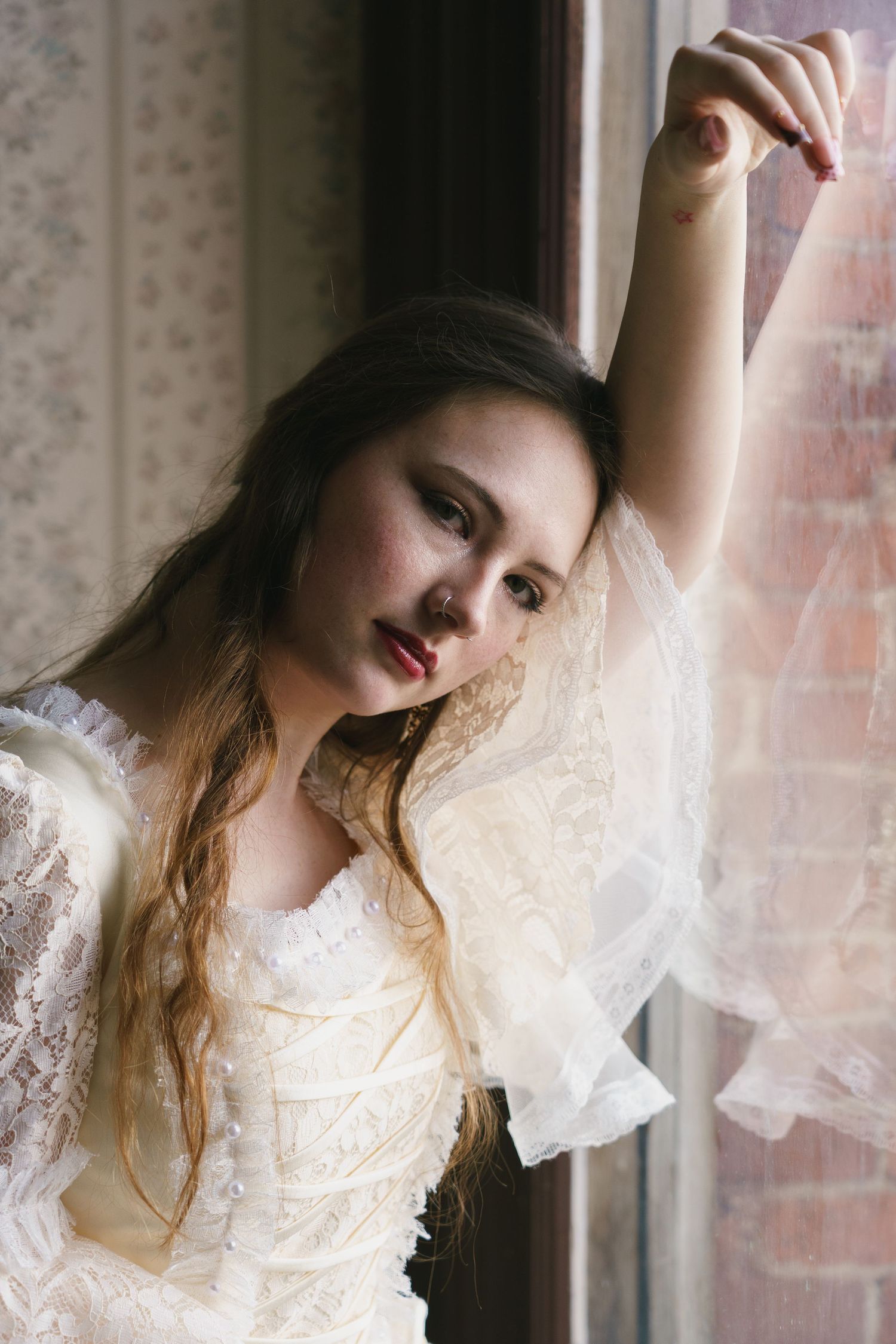 Young woman in vintage white lace dress poses gracefully by a window, holding sheer curtain with arm raised.