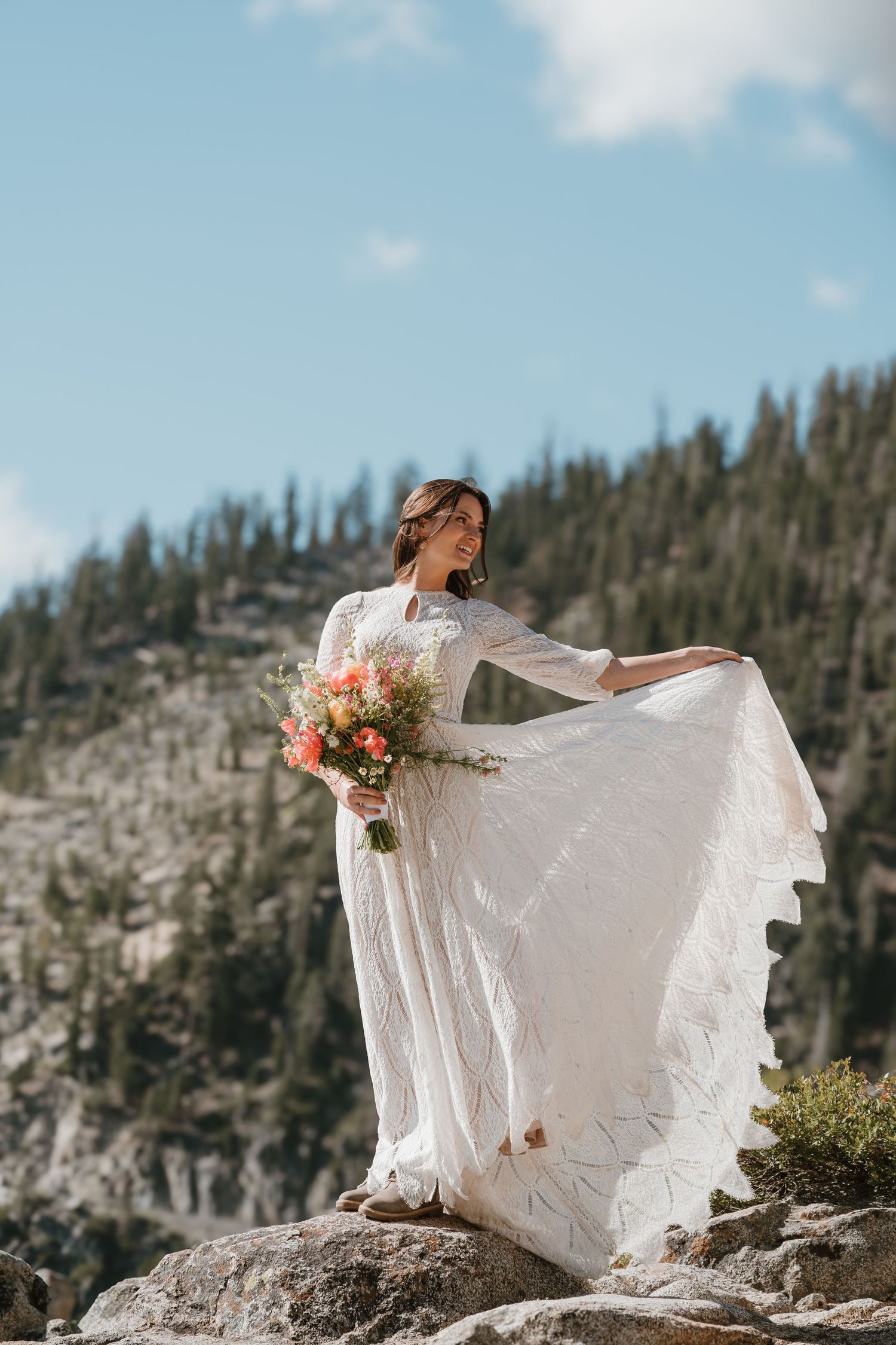 Bride in white lace gown holding coral bouquet on rocky mountain overlook with pine forest backdrop.