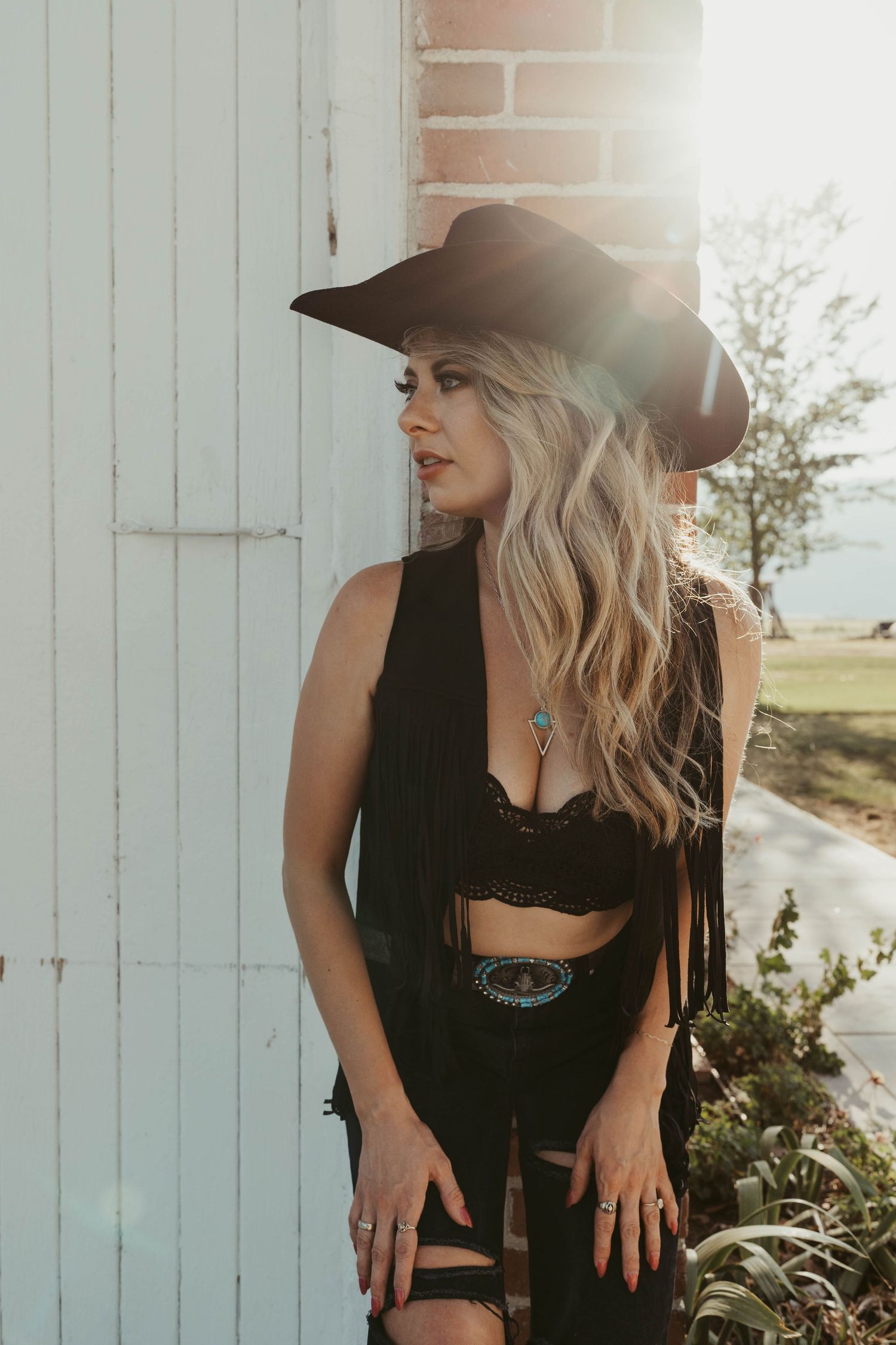 Country western styled portrait against white barn door featuring black outfit and hat with dramatic sun flare.