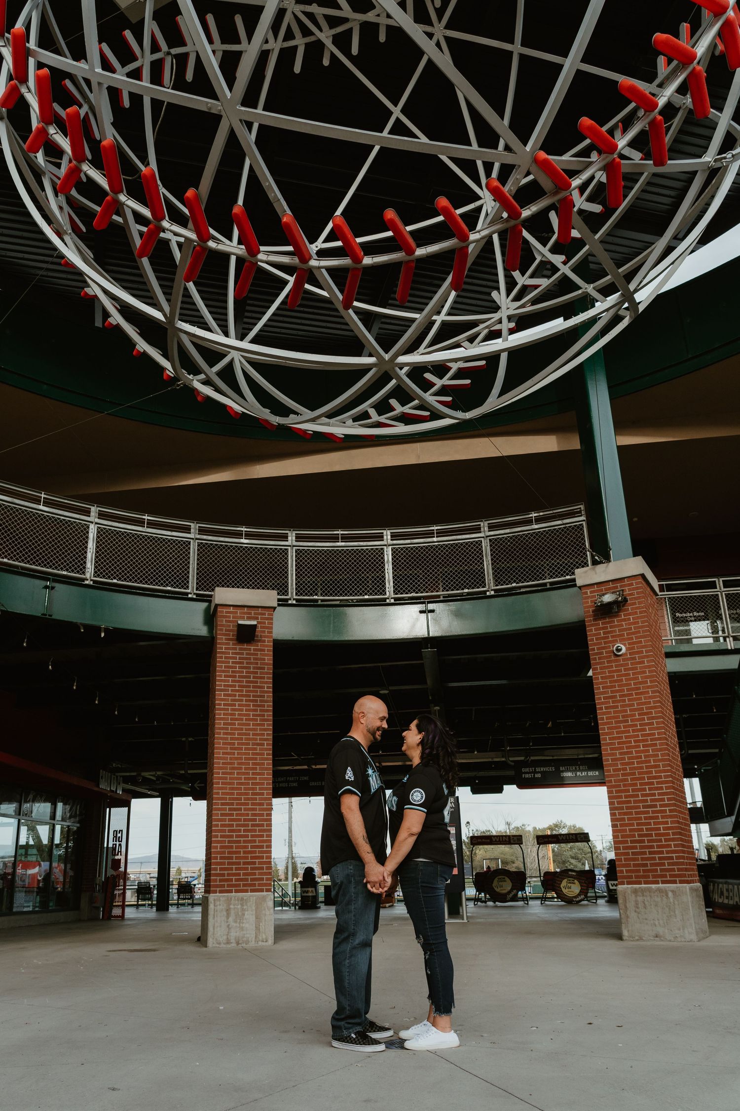 Couple holds hands under a giant baseball sculpture at a stadium entrance, both wearing matching black jerseys.