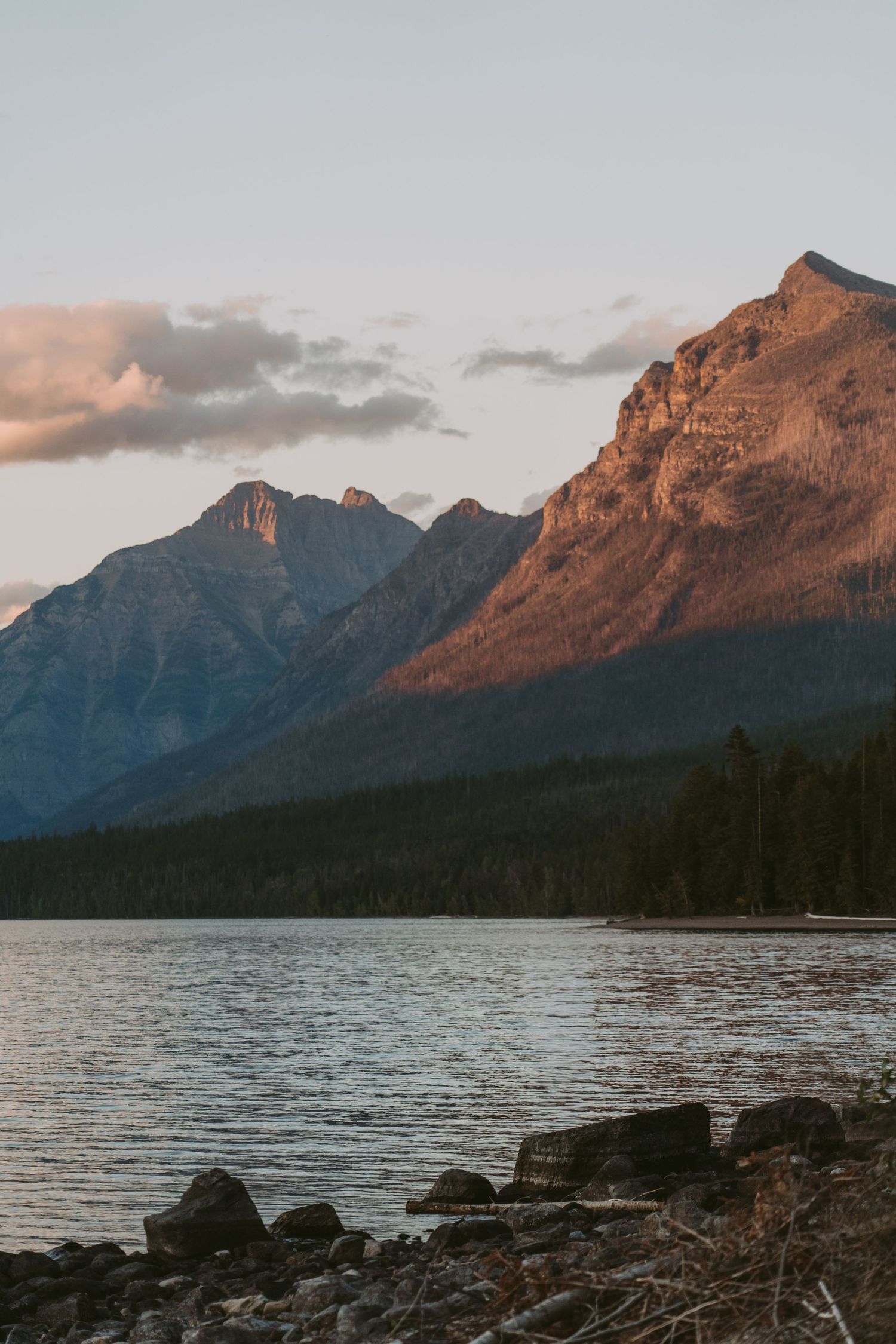 Rugged mountain peaks reflect golden sunlight over a serene lake with rocky shoreline at sunset in Banff National Park.