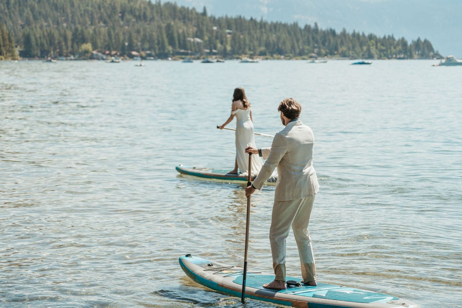 A couple paddleboarding together on a tranquil lake surrounded by forested hills on a sunny summer day.