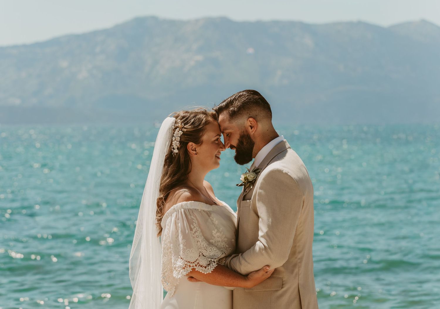 A romantic wedding couple embraces by turquoise waters with mountains in the background on their special day.