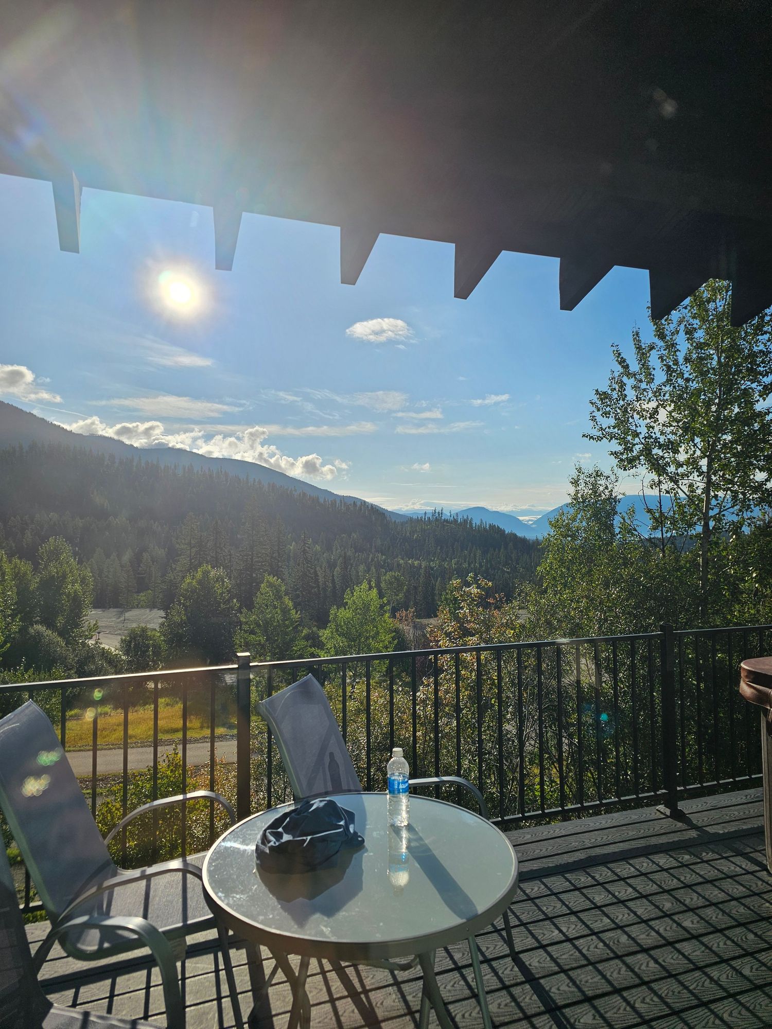 Mountain views from a deck with metal railing and outdoor seating against a backdrop of blue sky and evergreen forests.