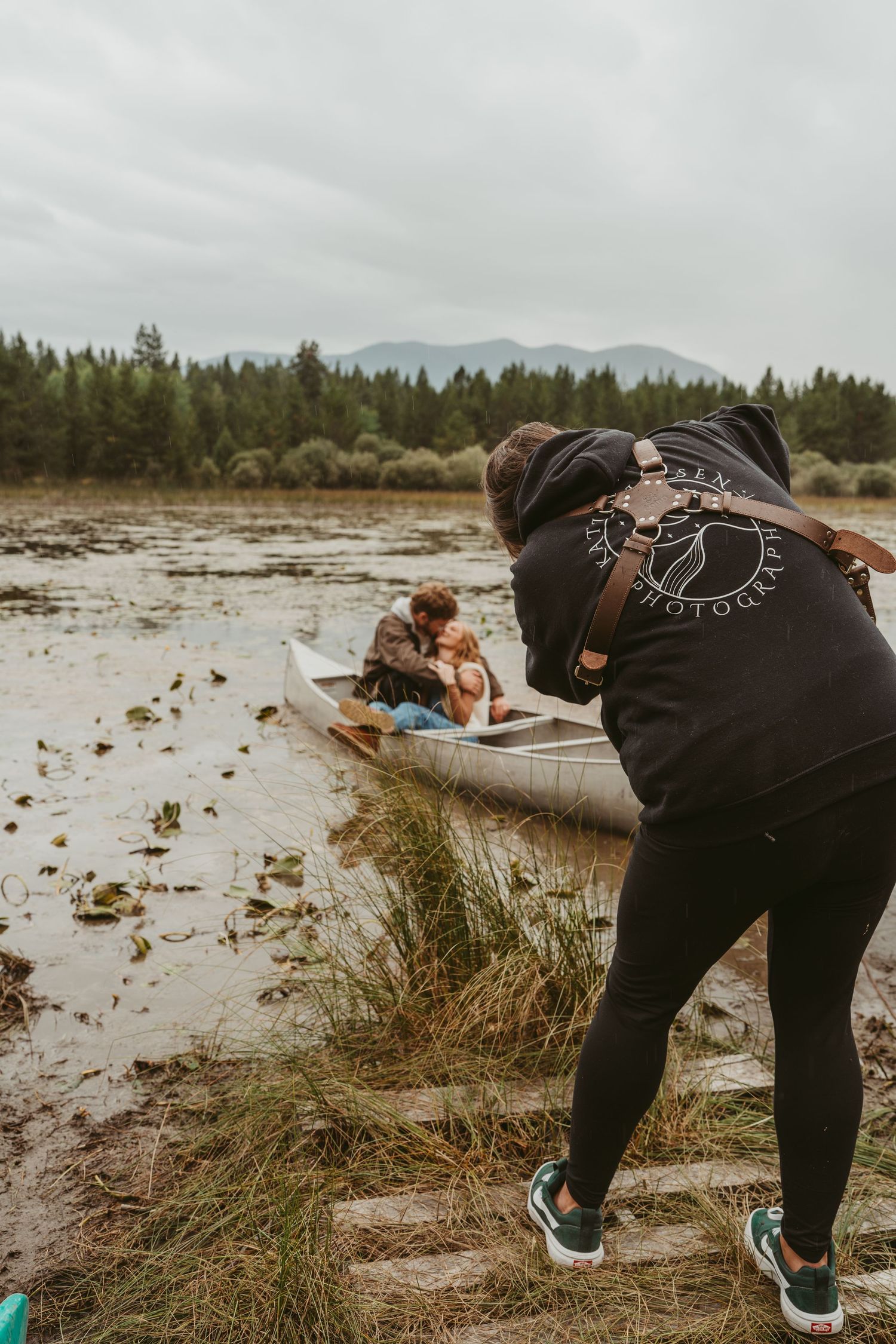 Someone in black leggings and green shoes bends down to photograph a couple sitting in a canoe nearby.