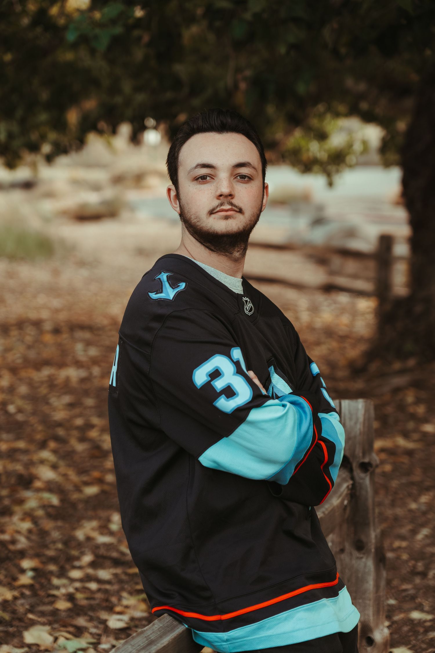 Person wearing black and turquoise jersey poses against autumn forest background with fallen leaves.