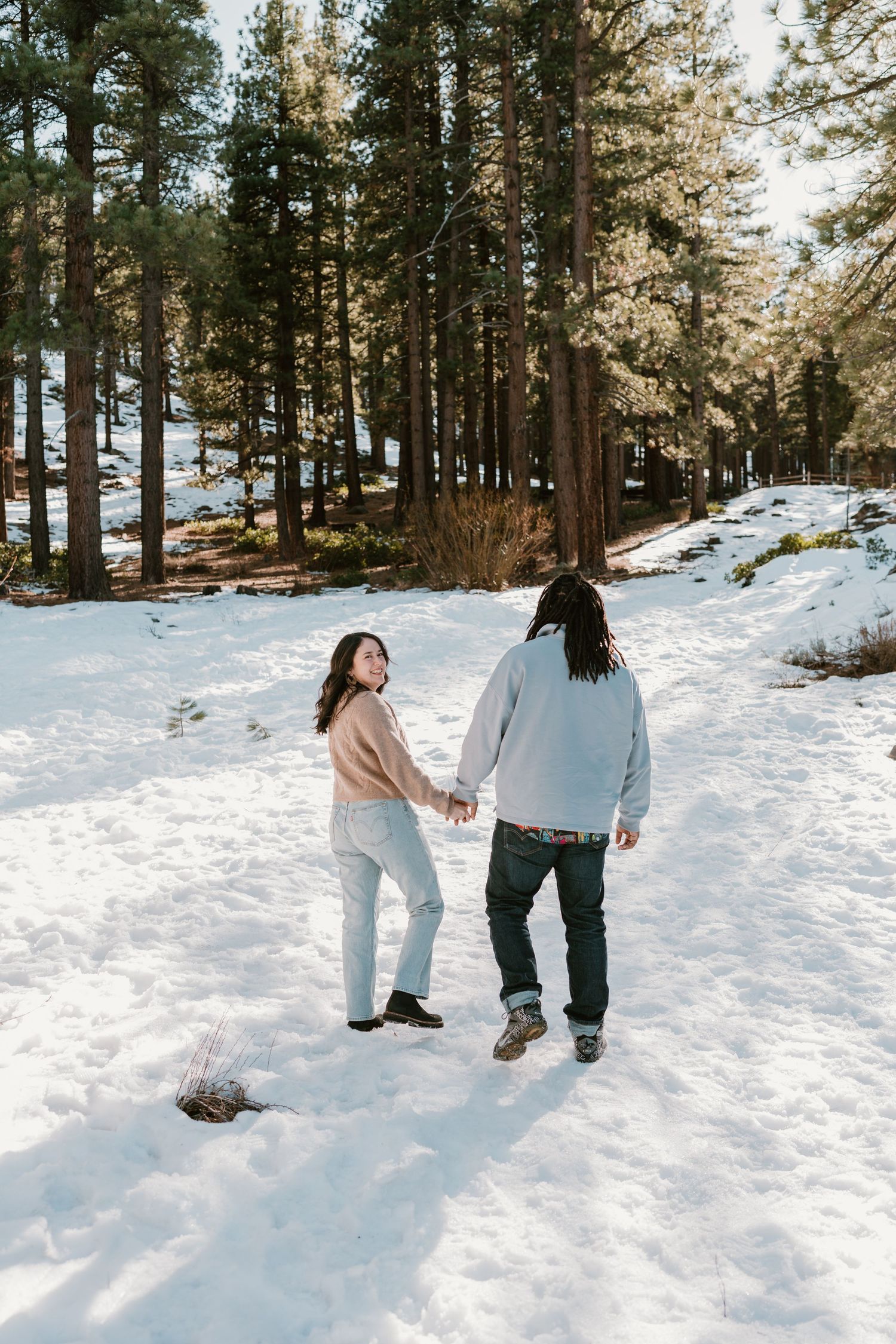 Couple holding hands walking through snowy pine forest, woman looking back smiling, winter outdoor scene.
