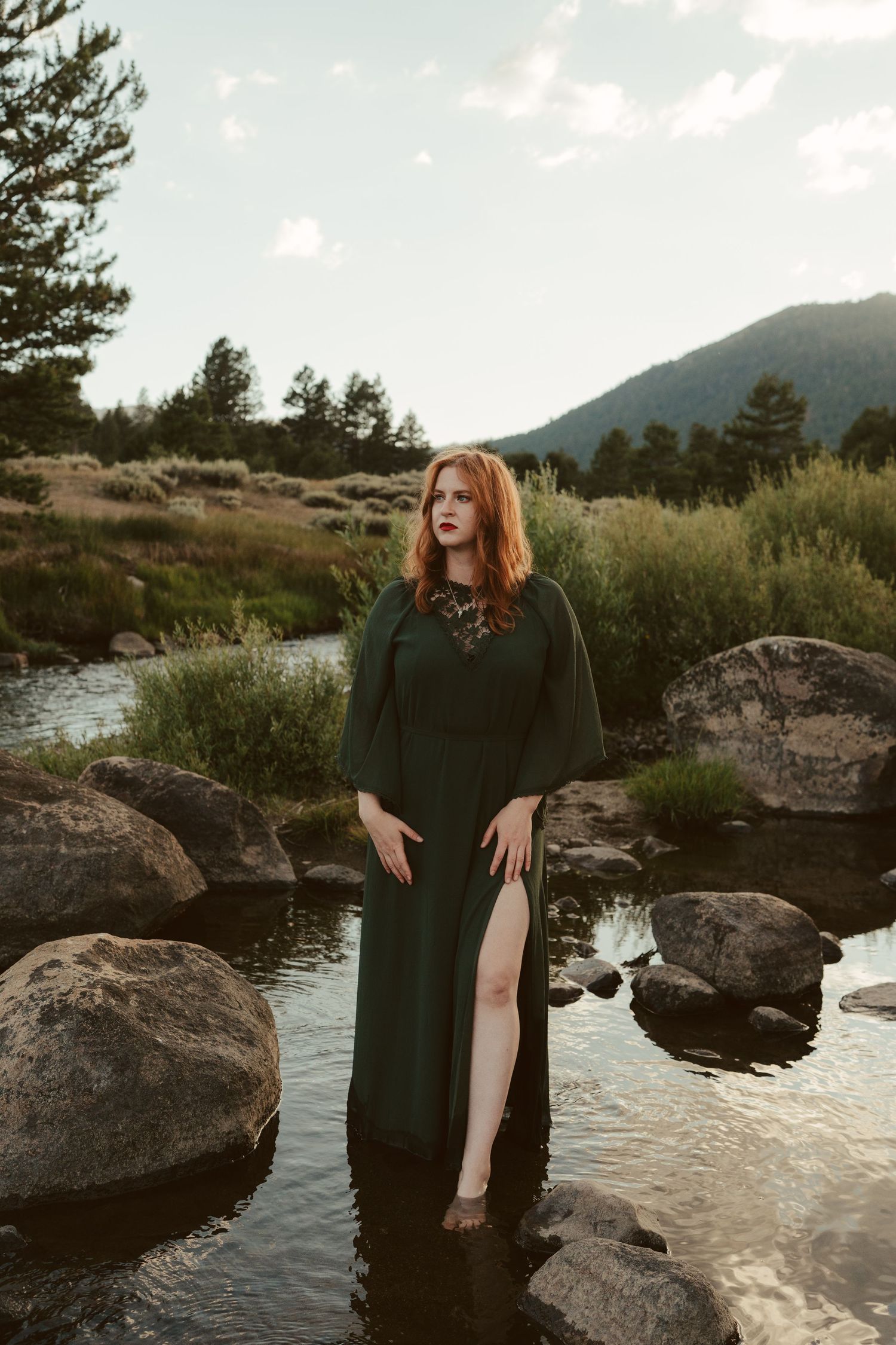Person in a green dress poses gracefully on rocky riverbank with mountains and evergreen trees in background at sunset. Hope Valley, CA