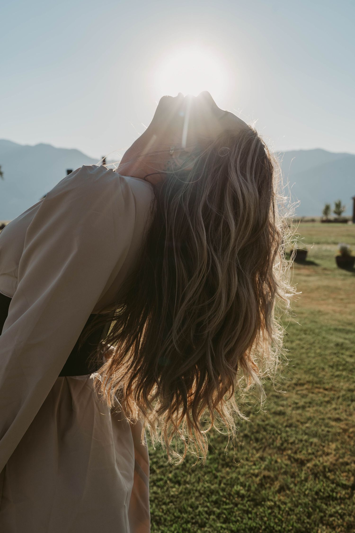 Person with flowing hair stands in a grassy field at sunset with mountains in the background.