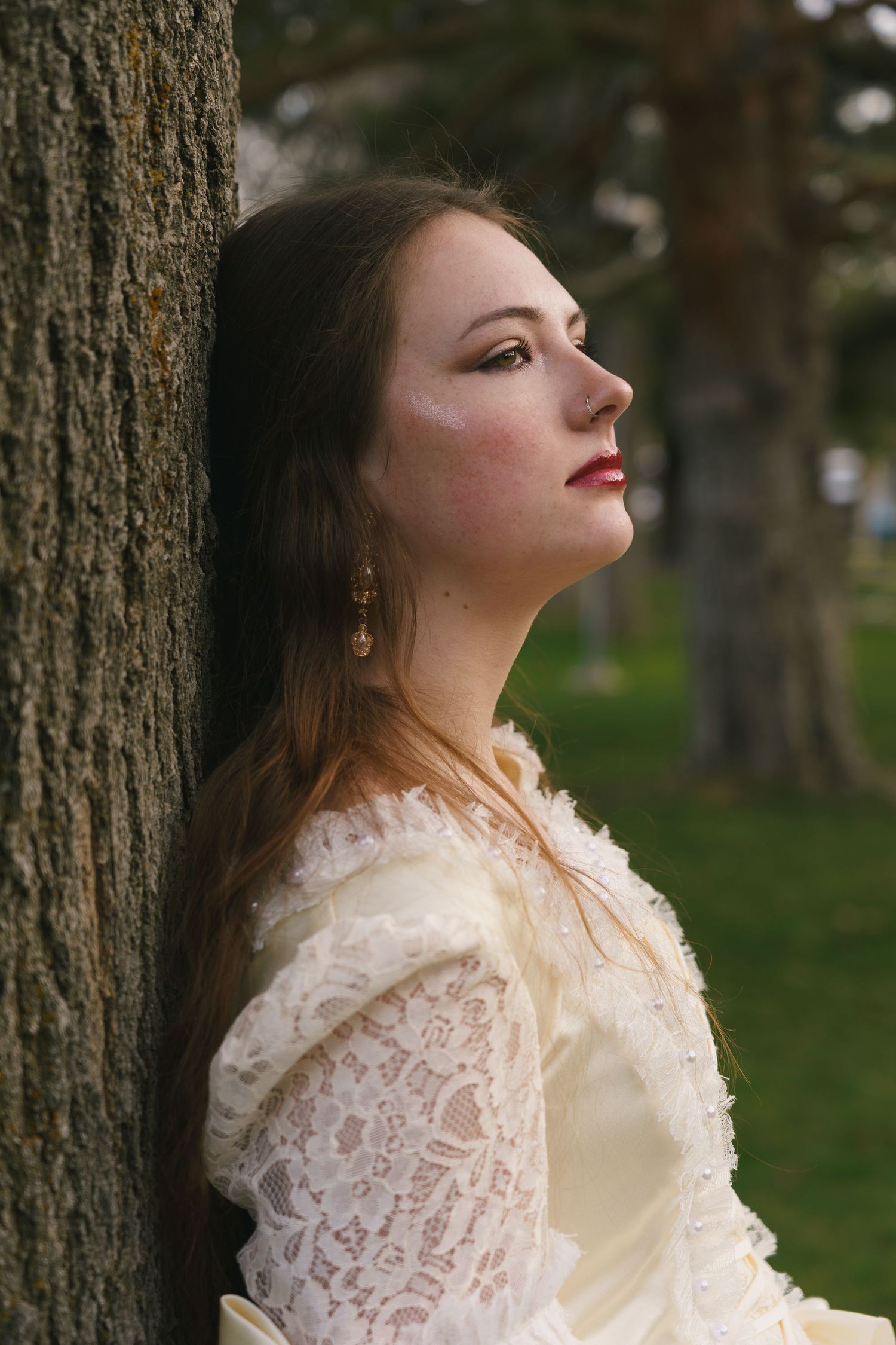 Young woman in Victorian lace dress leaning against tree trunk outdoors, gazing upward thoughtfully.