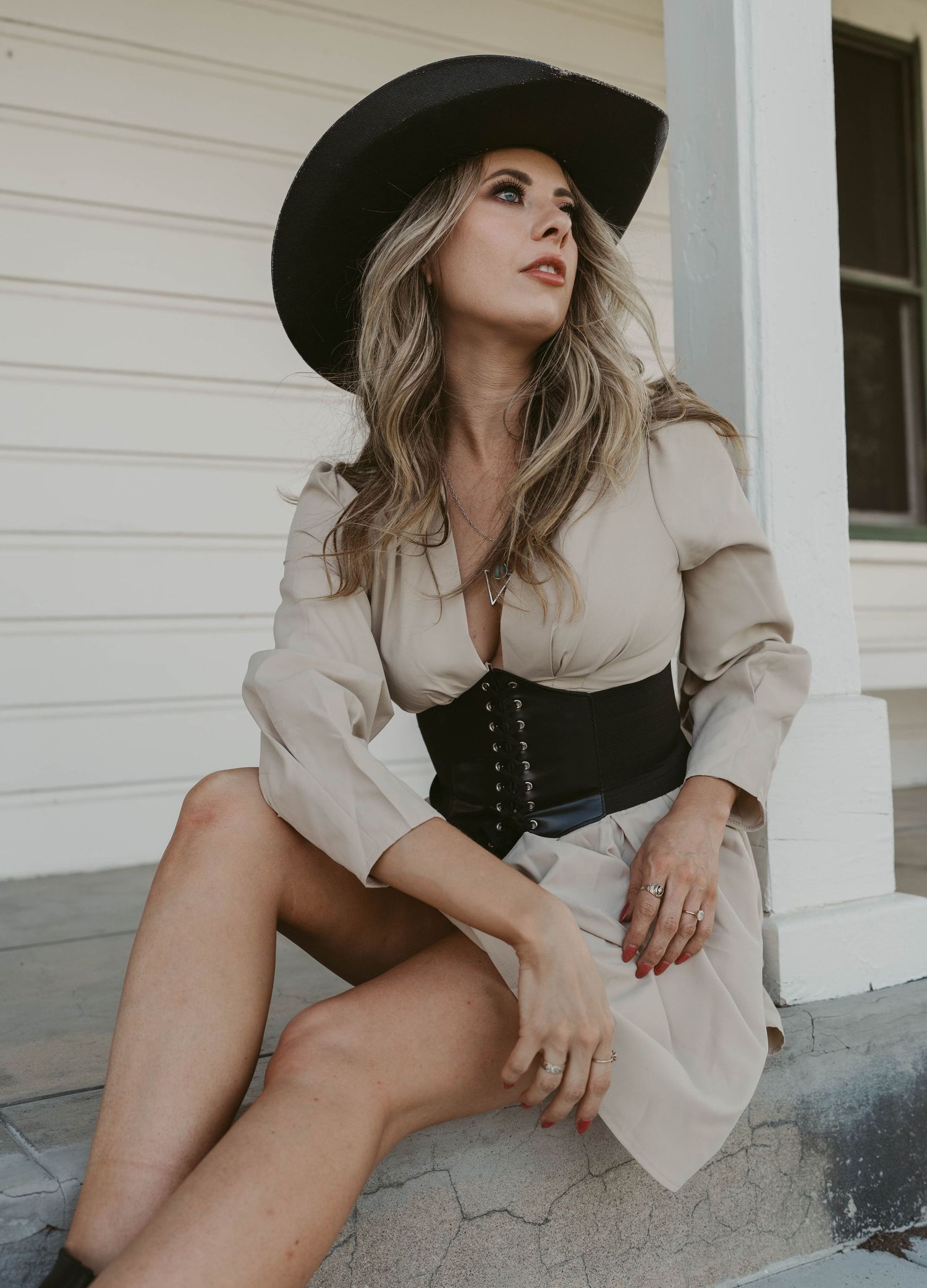Stylish portrait in beige dress with black corset belt and wide brim hat against white wood siding.