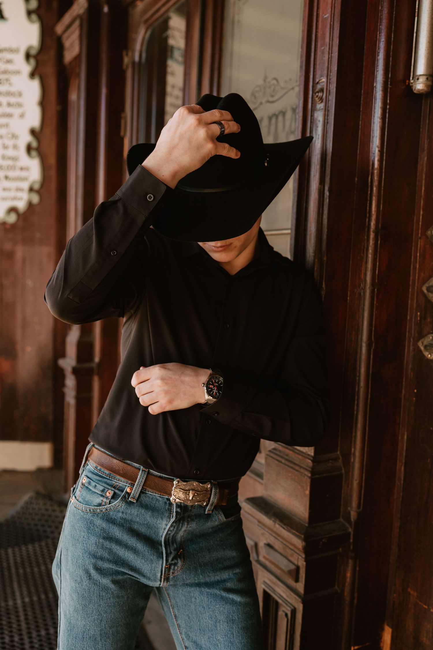 Figure in black turtleneck and blue jeans poses against dark wooden doorway.