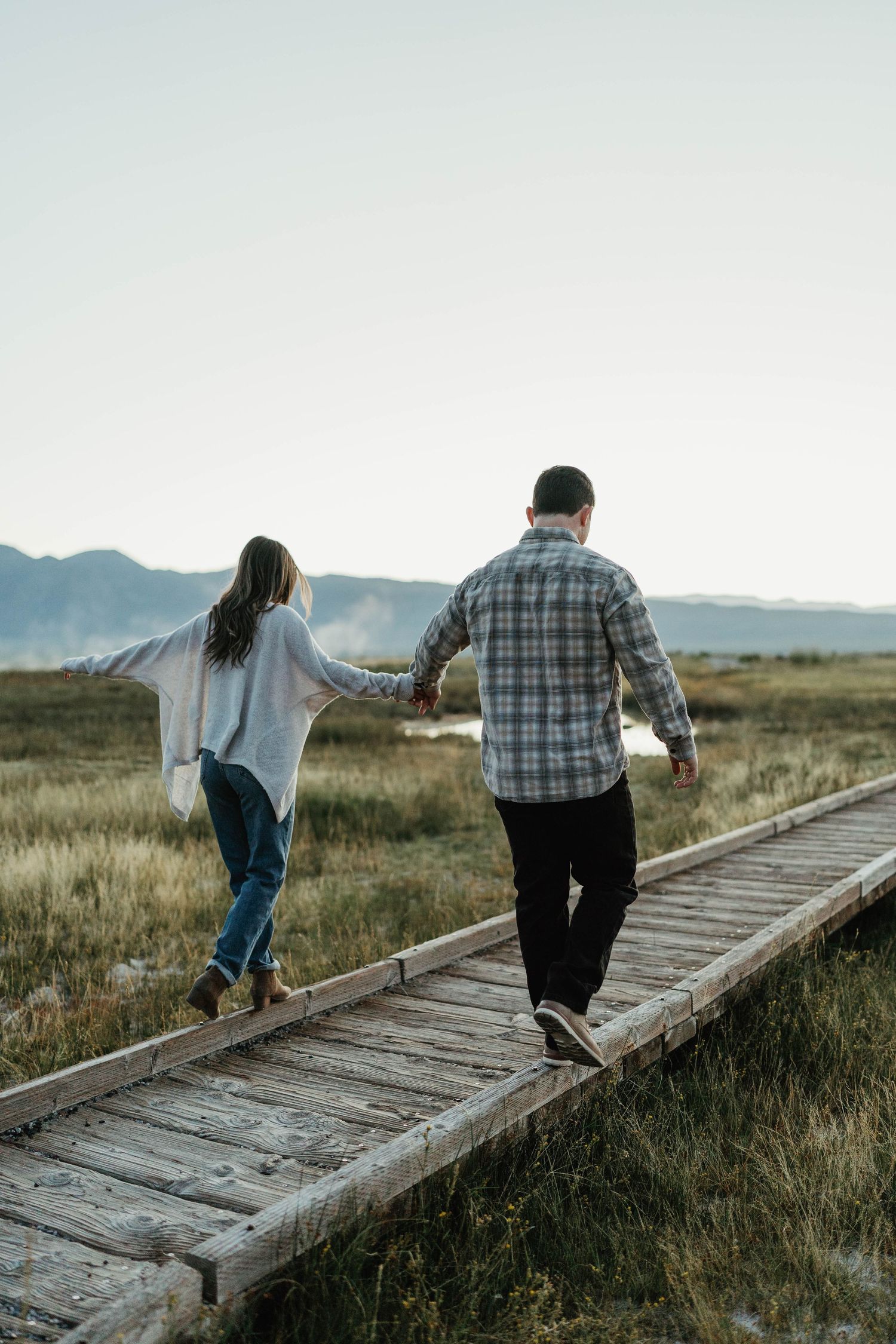 Two people walk along a wooden boardwalk path through a grassy field with mountains in the distance.