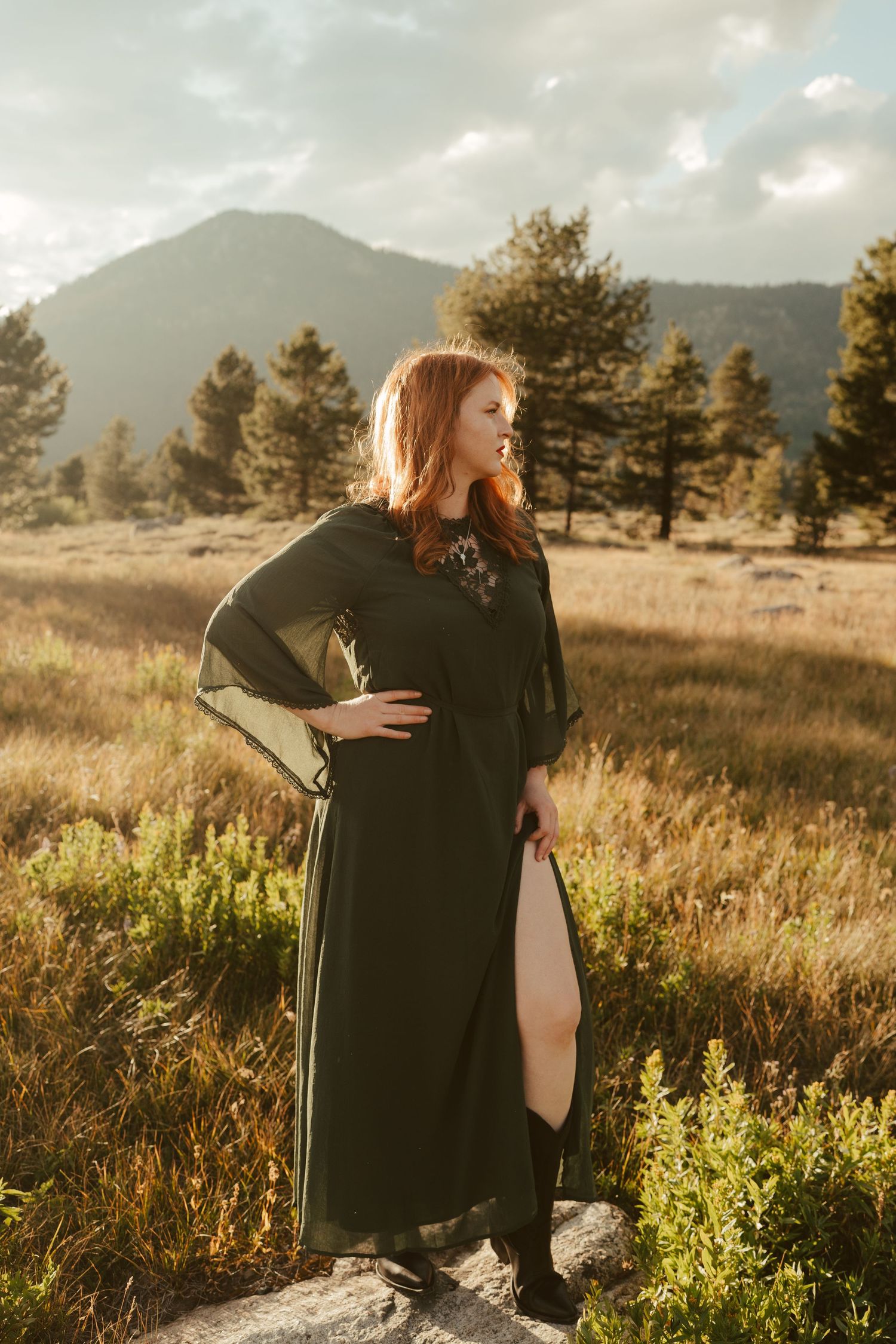 Red-haired woman in dark green maxi dress poses in mountain meadow with pine trees and cloudy sky backdrop.