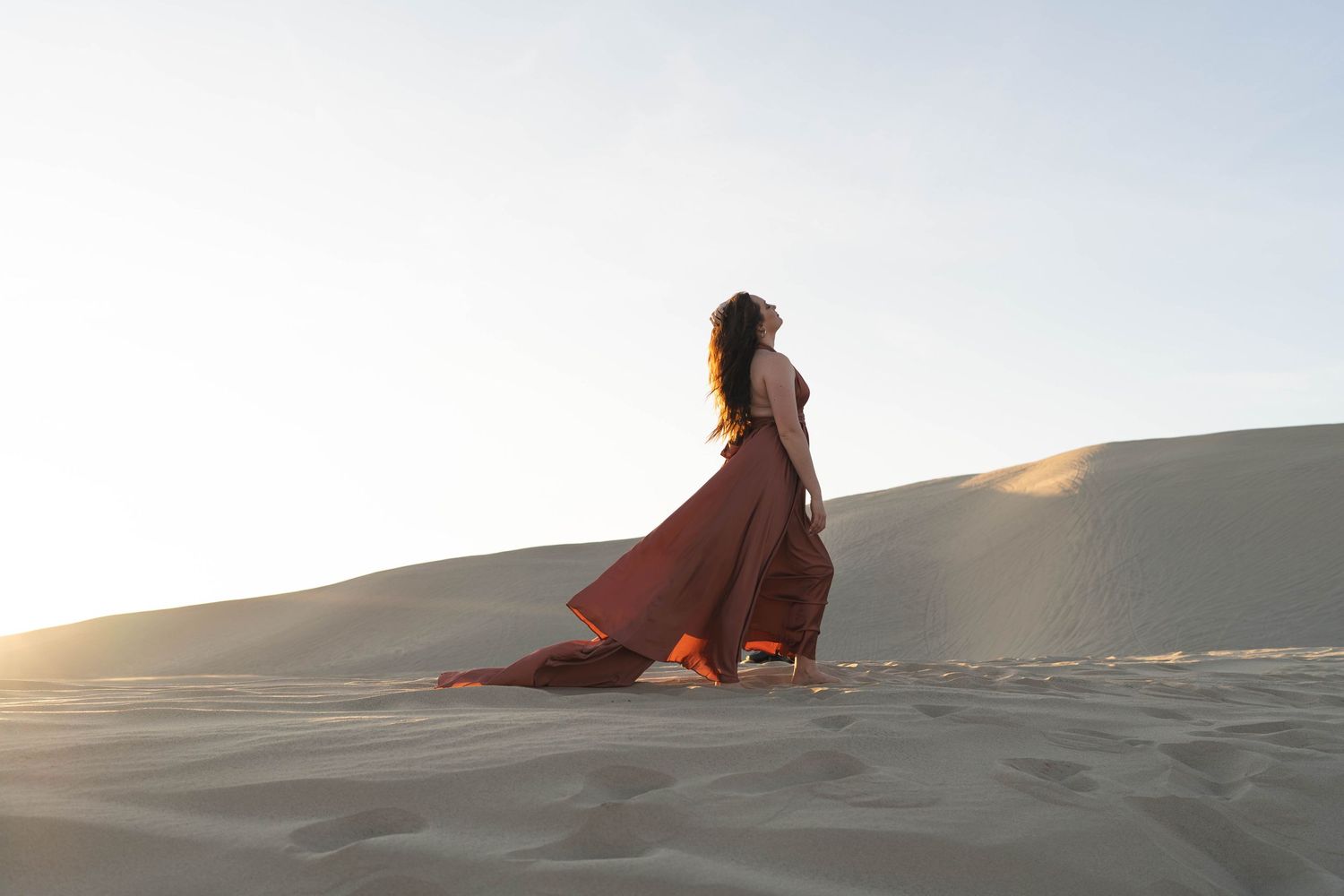 A flowing coral dress catches the wind as a person stands atop golden sand dunes at sunset.