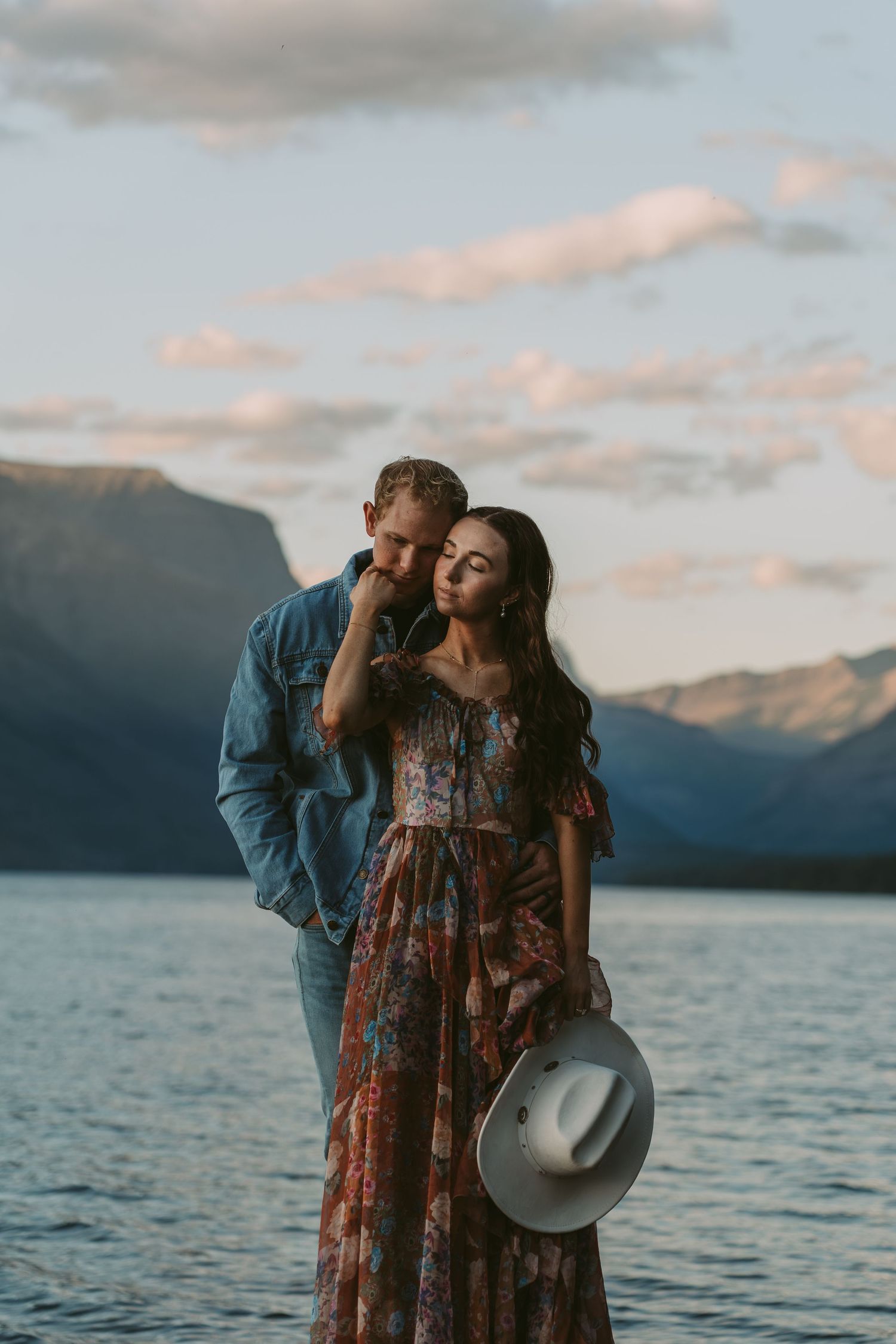 A couple shares an intimate moment by a scenic mountain lake at sunset while holding a cowboy hat.