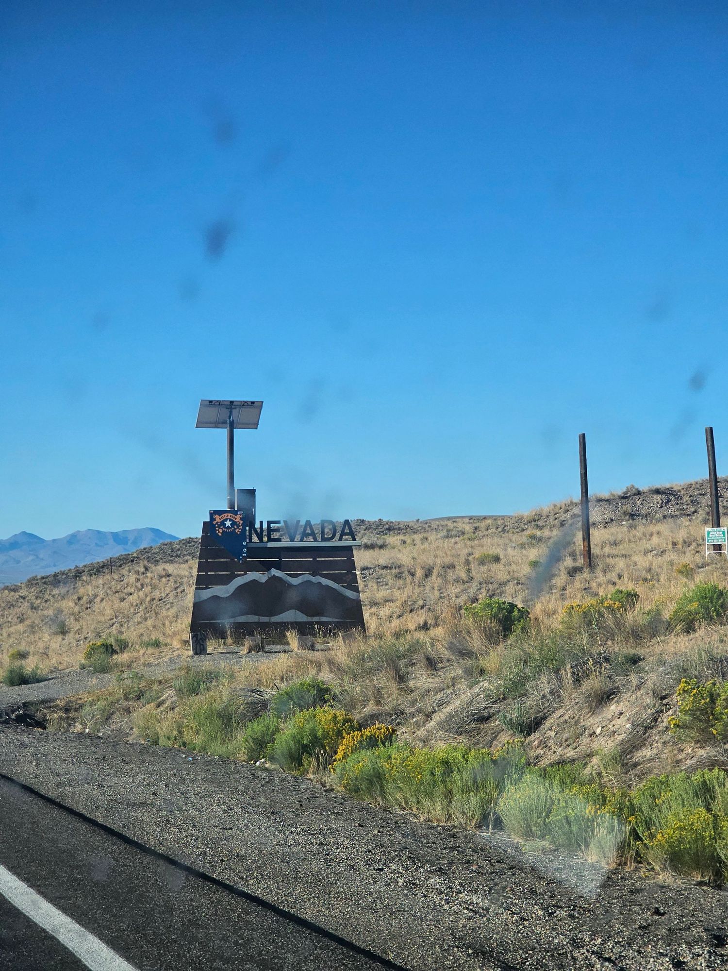 A Nevada state border sign stands along a desert highway with mountains and blue sky in the background.