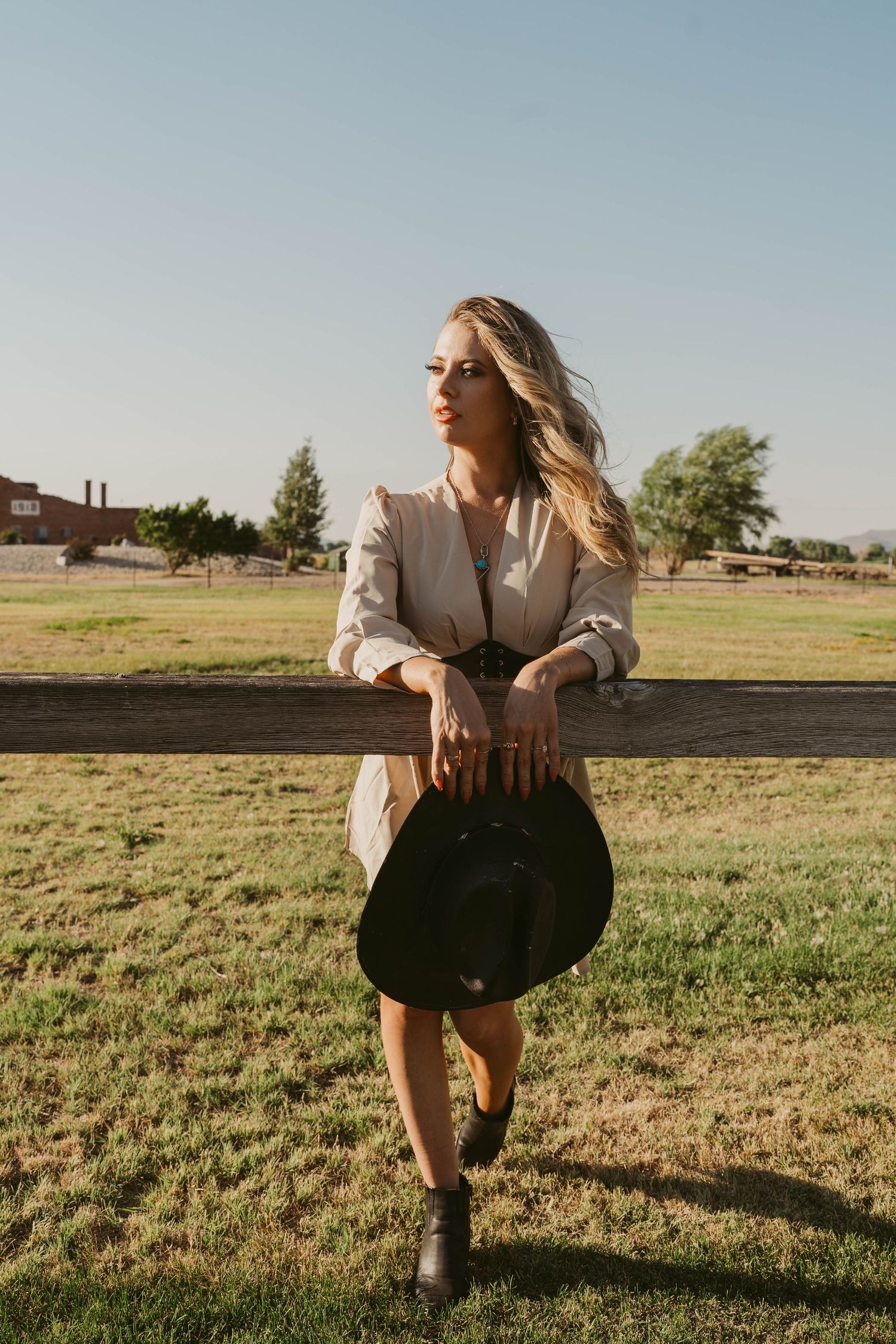 A person in a beige dress holding a black cowboy hat leans against a wooden fence in a grassy field at sunset.