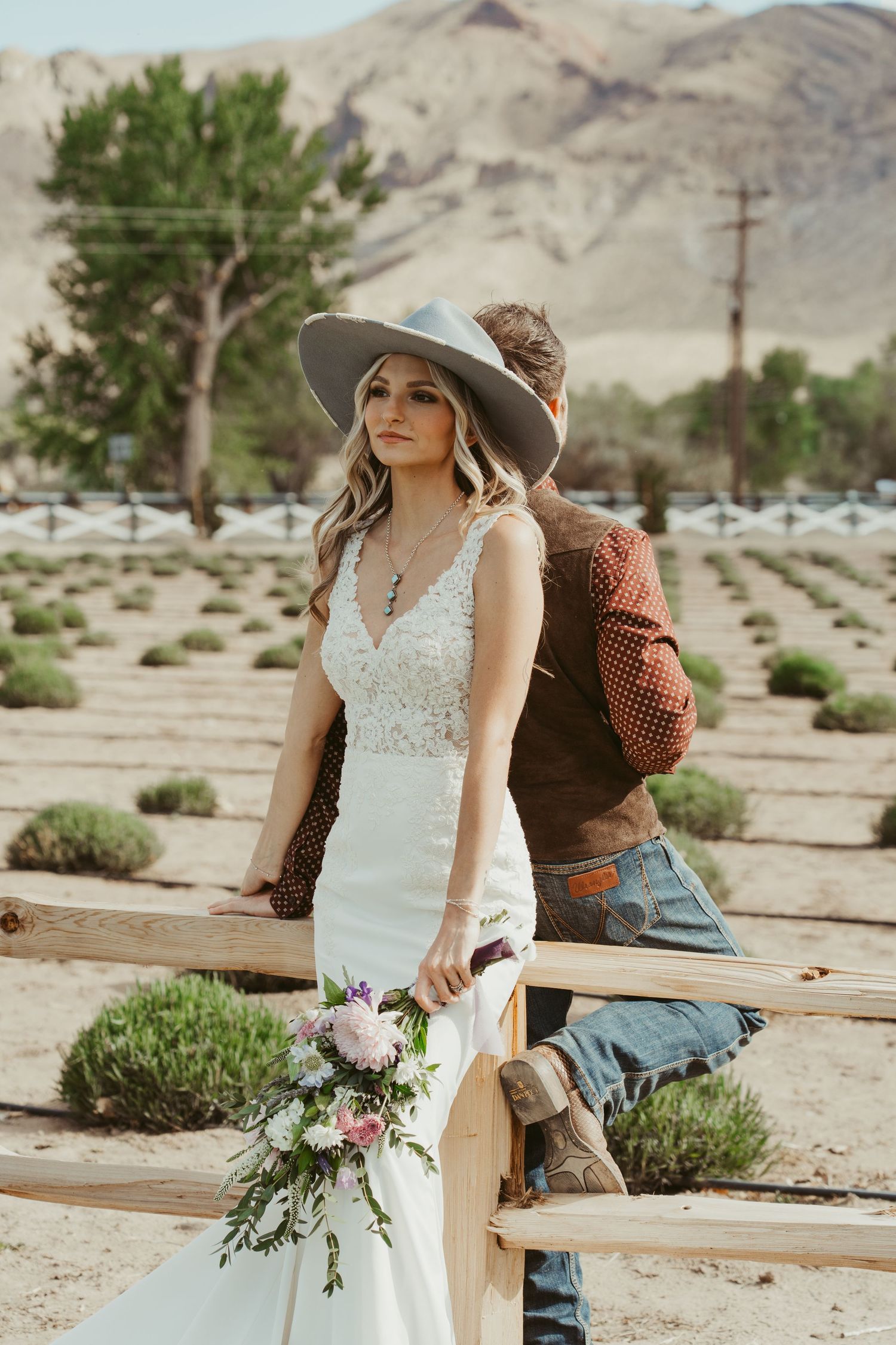 Bride in white lace gown and wide-brim hat holds floral bouquet by a fence with desert mountains behind.
