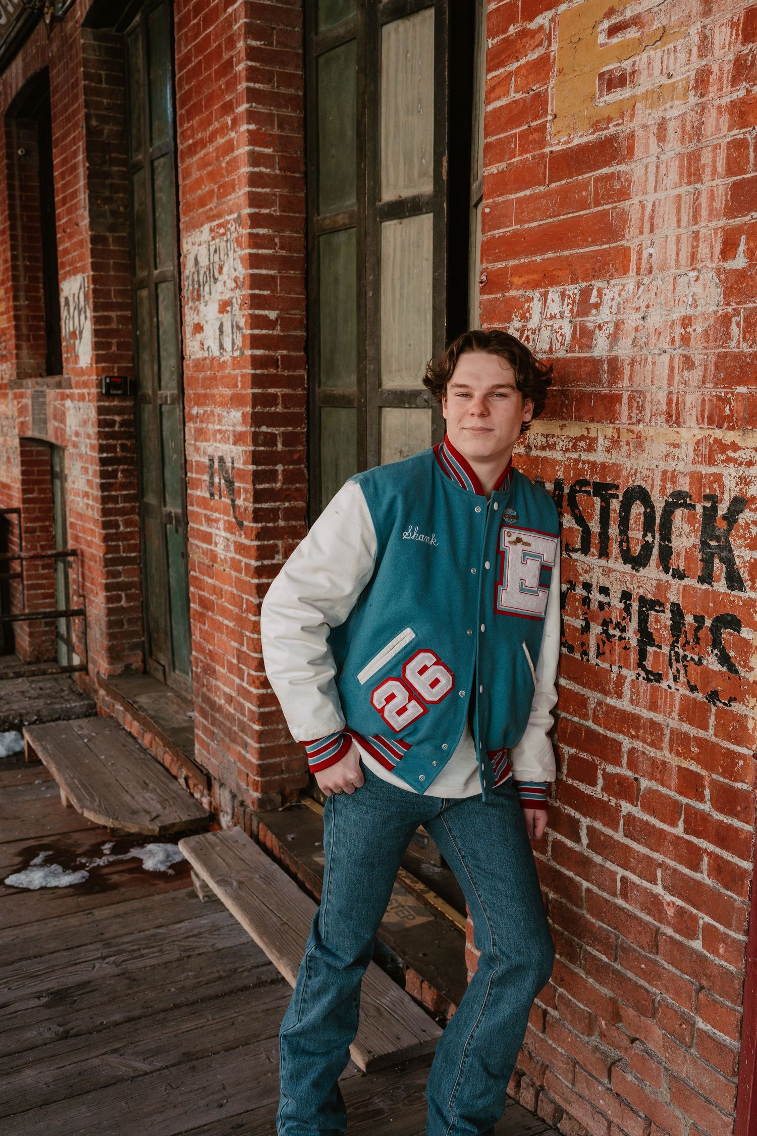 Person wearing a varsity jacket and jeans poses against rustic brick wall in casual style photoshoot.