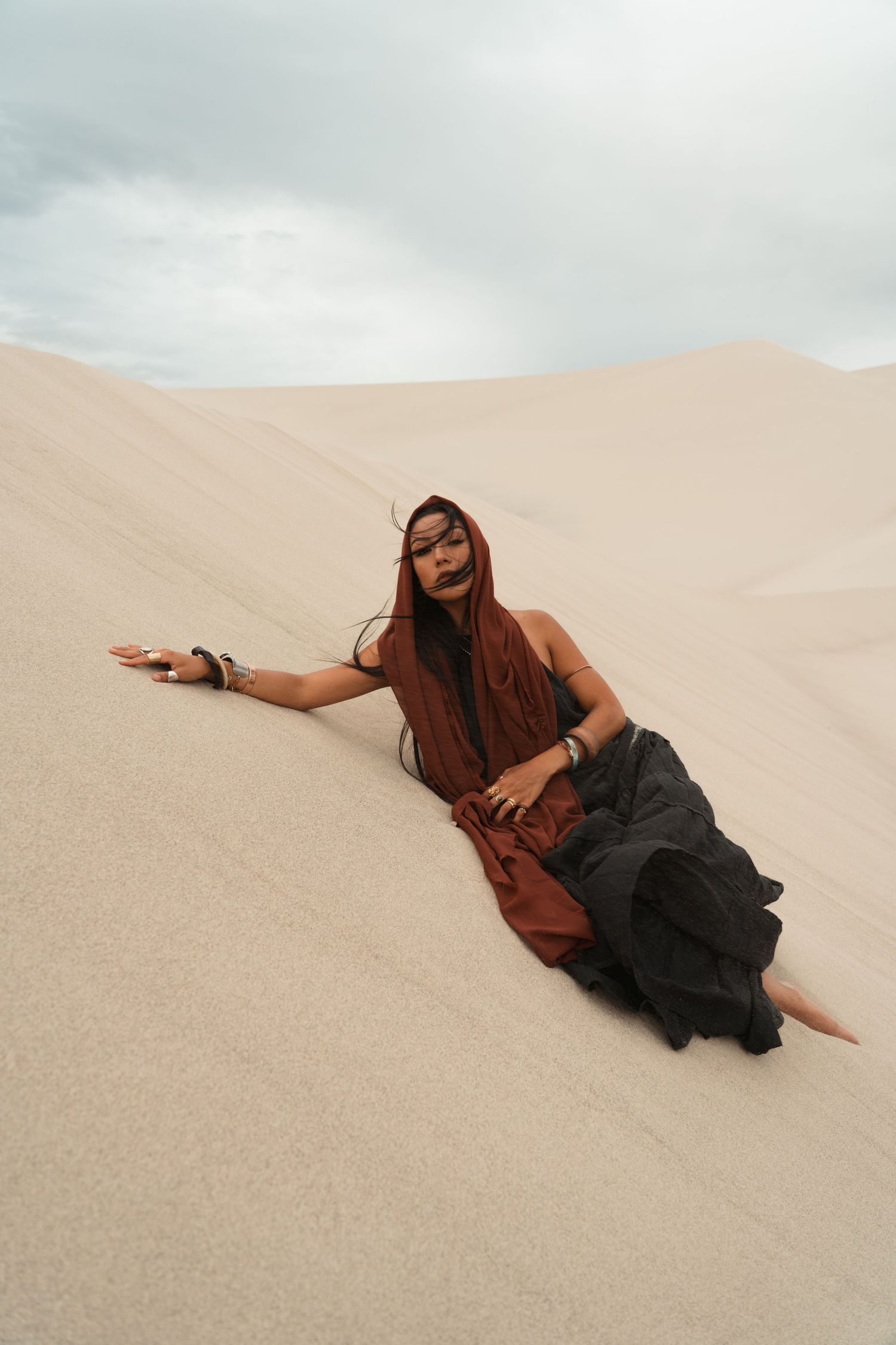 A woman in rust and black desert attire reclines gracefully on sandy dunes under a cloudy sky.