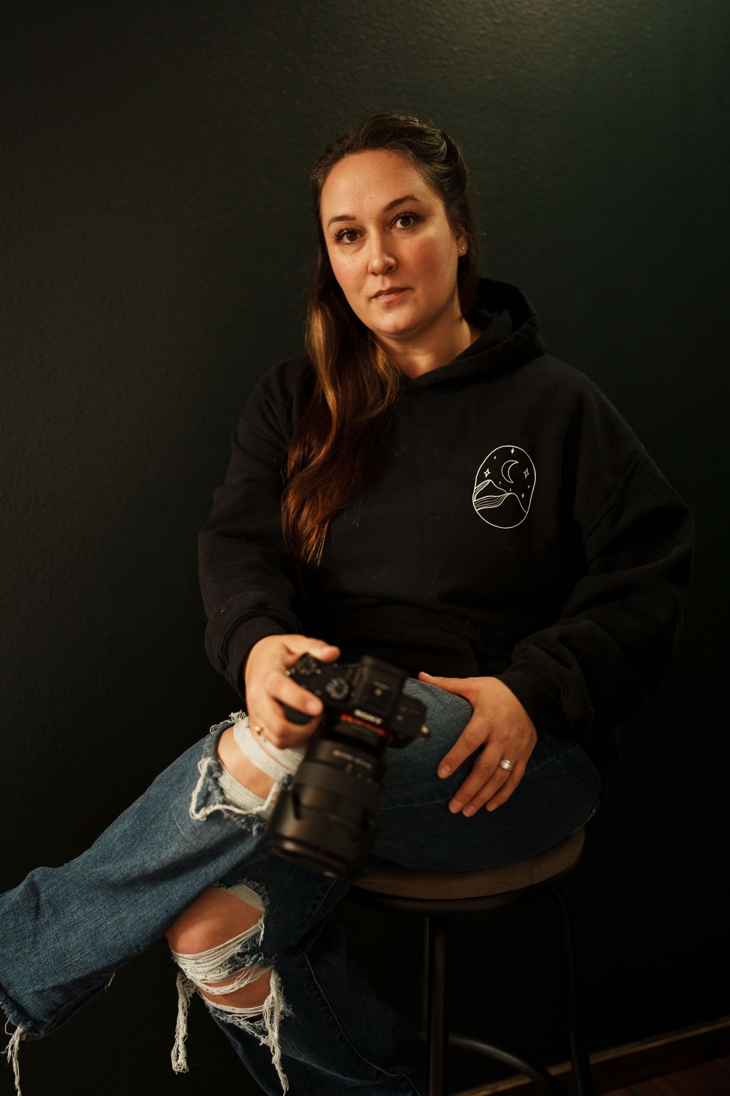 A photographer in a black hoodie and ripped jeans holds a camera while seated against a dark background.