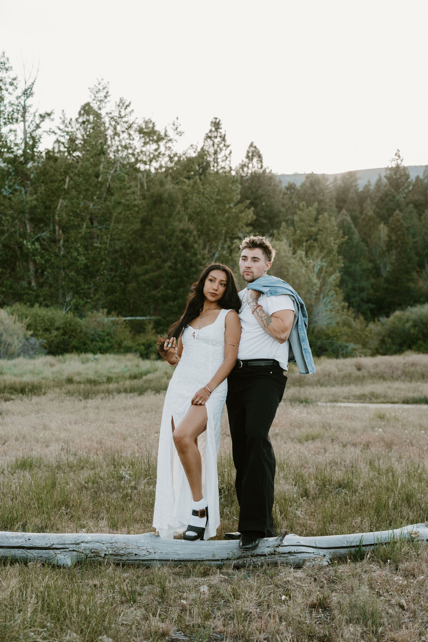 Couple posing outdoors in a field, woman in white dress and man in white shirt with jacket over shoulder.