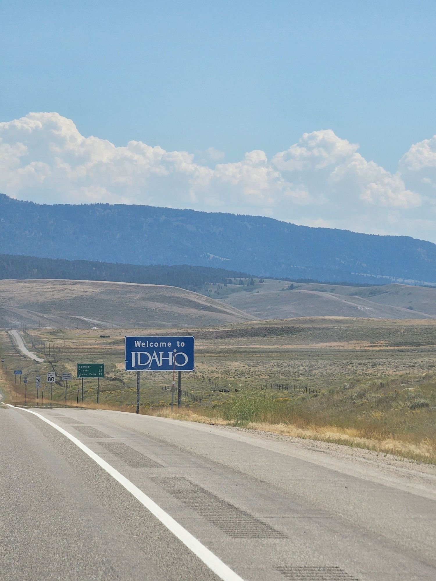 A highway road leads to the 'Welcome to Idaho' sign with rolling hills and mountains visible in the distance under blue skies.